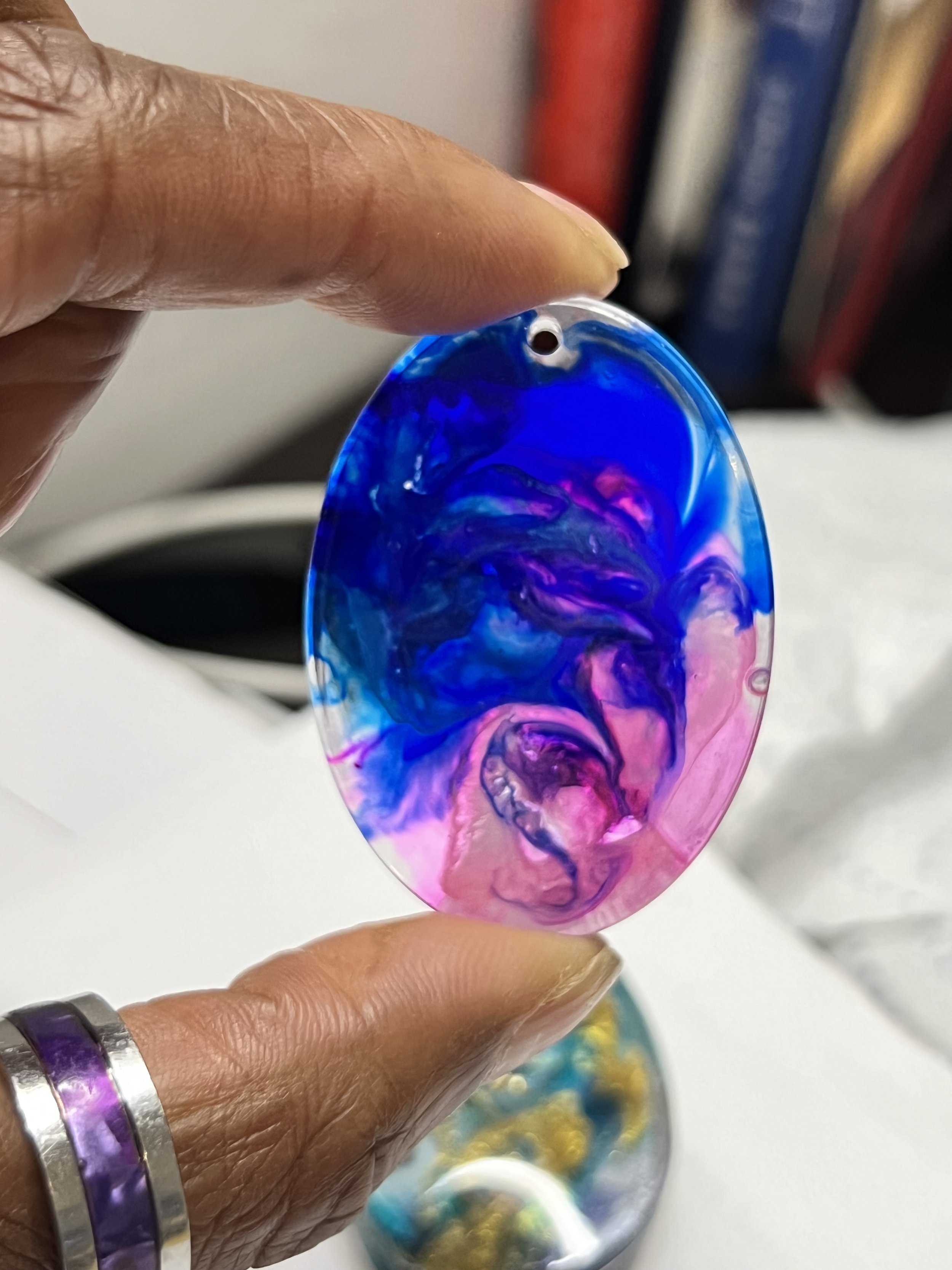 A colorful, oval-shaped glass pendant with a swirling pattern of blue, pink, and purple hues, held between a person's fingers.