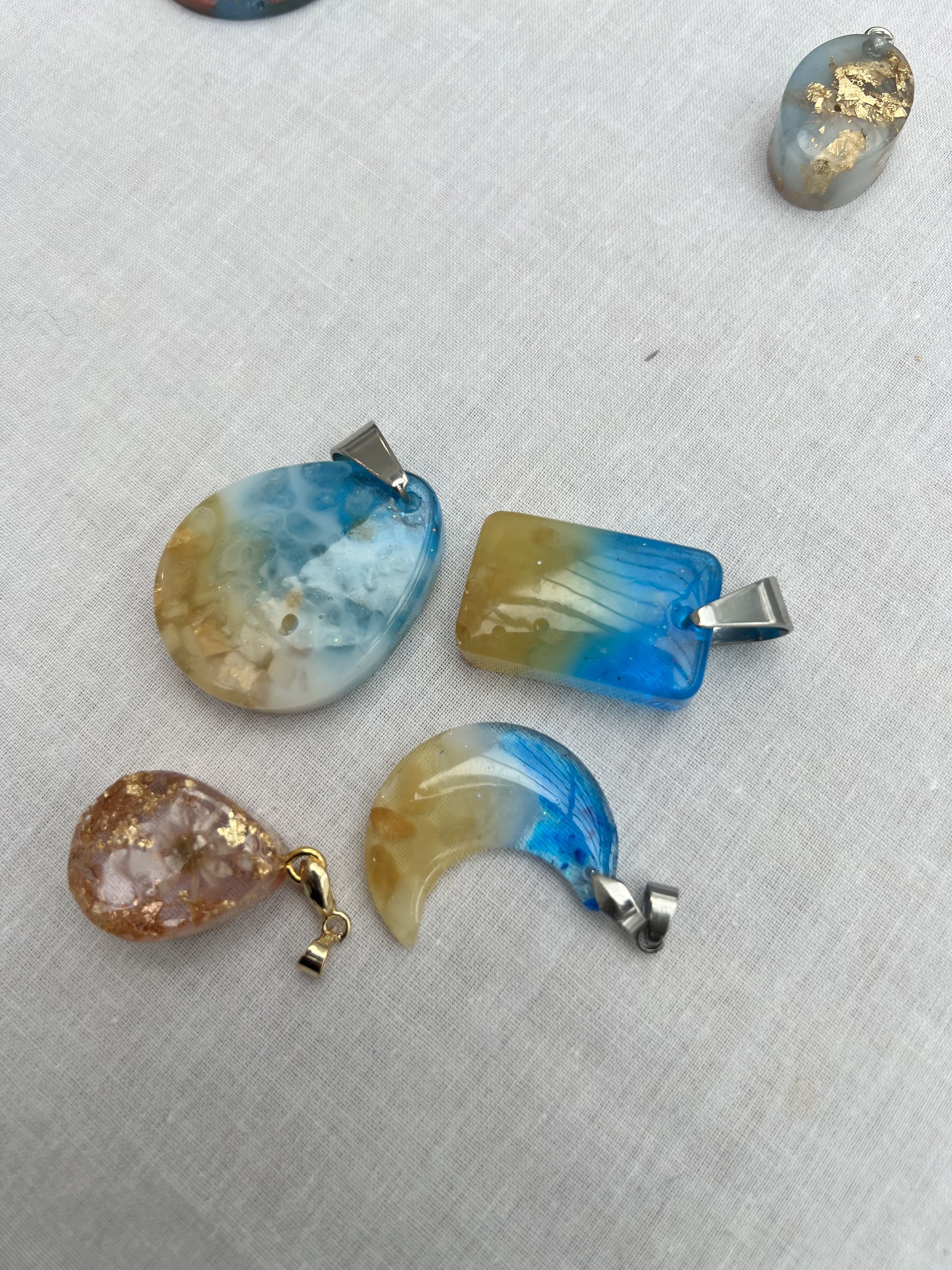 Five resin pendants with silver or gold hooks on a white fabric surface, featuring blue, gold, and white colors, and one small smooth stone with gold accents.