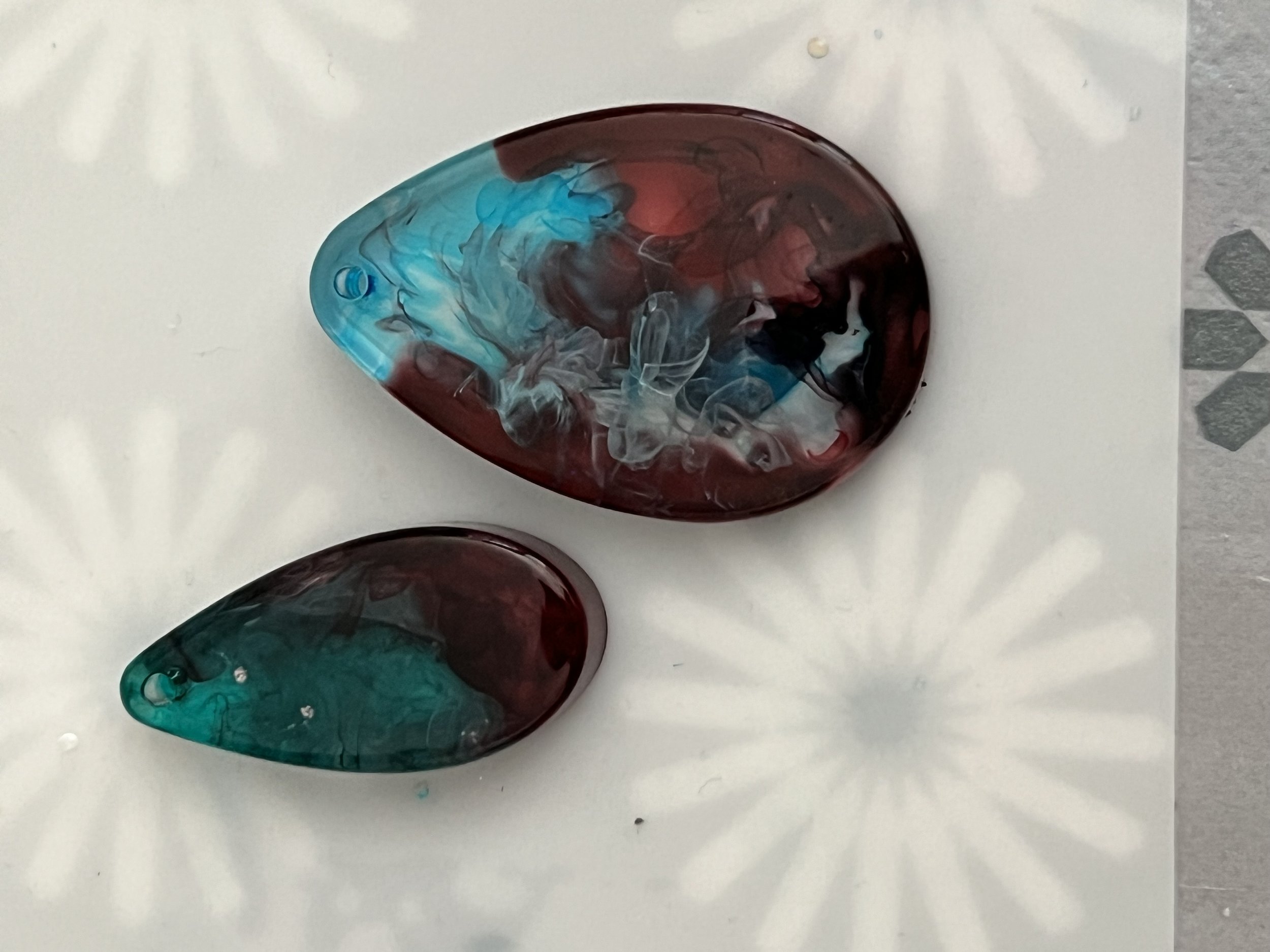 Two teardrop-shaped glass pieces with swirling red, black, blue, and white colors, resting on a white surface with faint floral patterns.