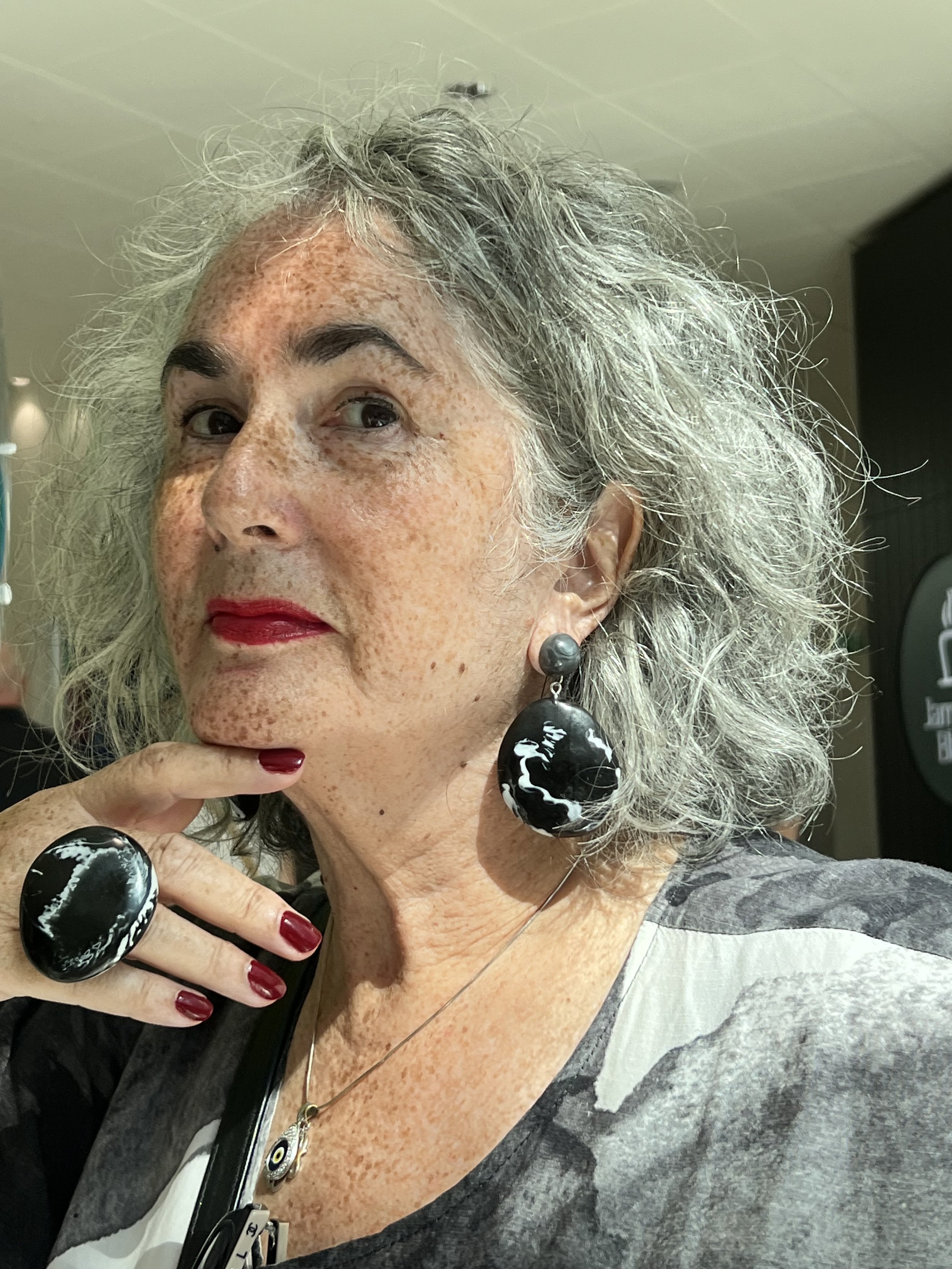 A mature woman with curly gray hair, freckles, and red lipstick, wearing large black and white marble earrings, a matching ring, a black and gray top, and a necklace with an eye charm, posing with her hand near her face.