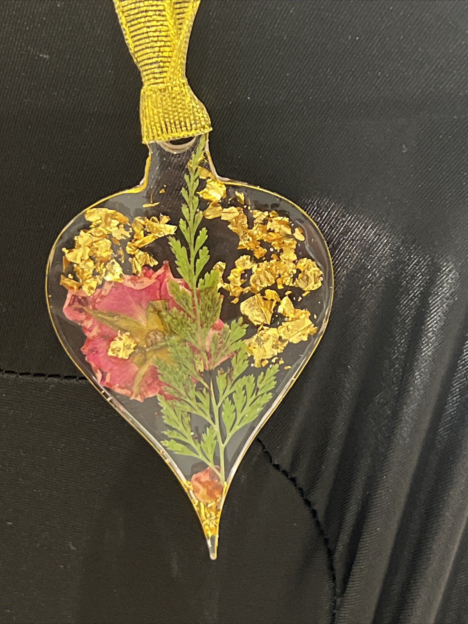 A heart-shaped glass ornament containing a small plant, yellow flower fragments, and a pink flower, hanging from a yellow ribbon.