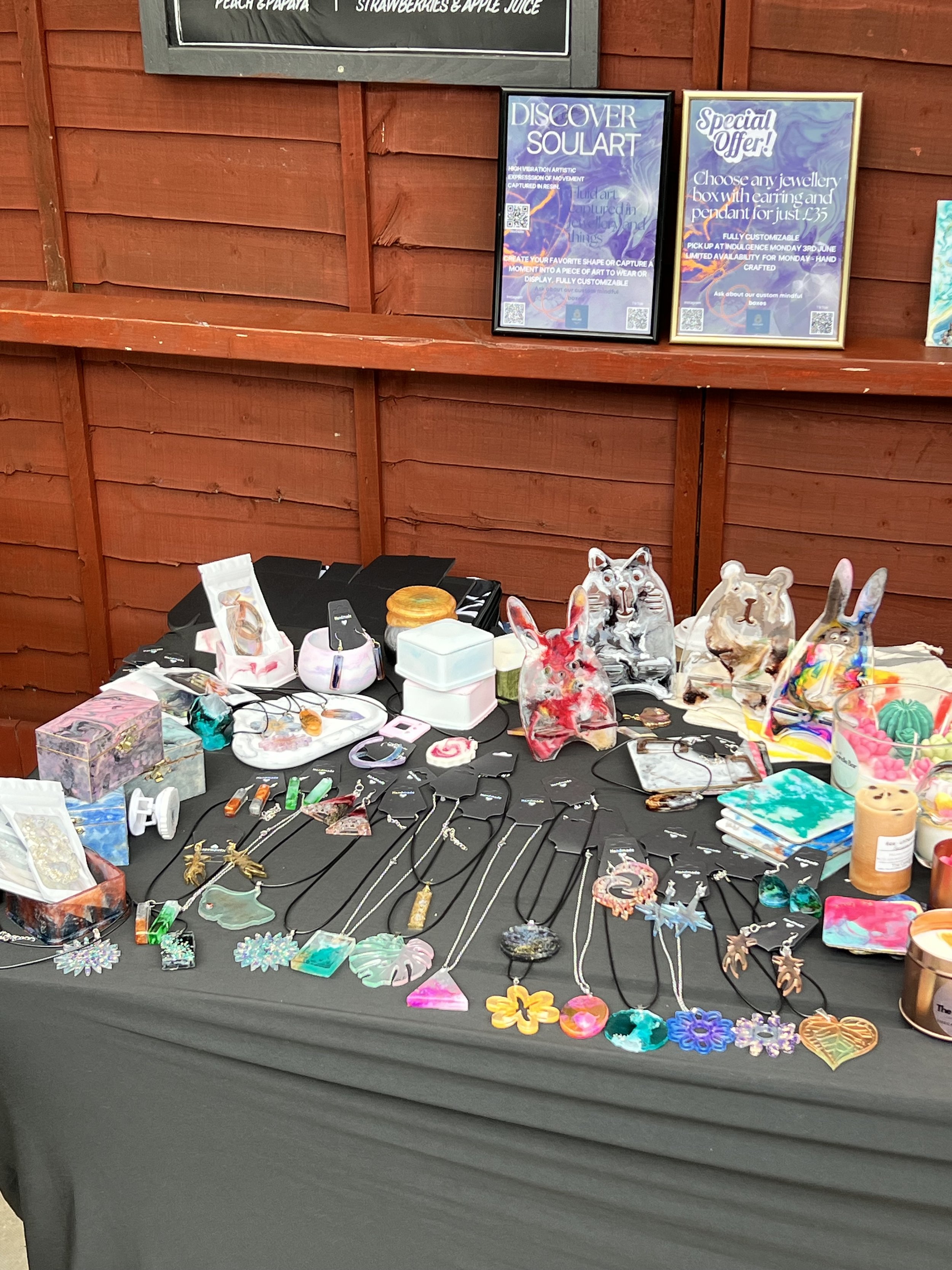 Display table with colorful necklaces, jewelry, and art pieces, alongside small trinkets and decorative items, set against a wood-paneled wall.