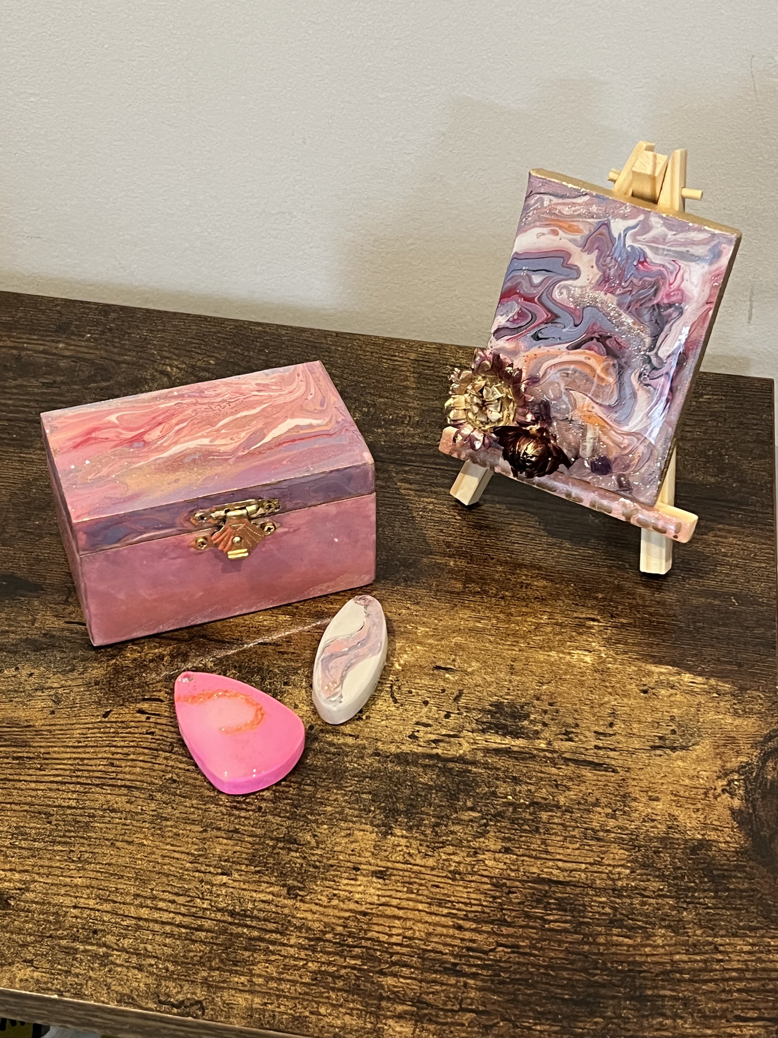 A pink jewelry box, two polished stones, and a small easel with a colorful abstract painting on a wooden surface.