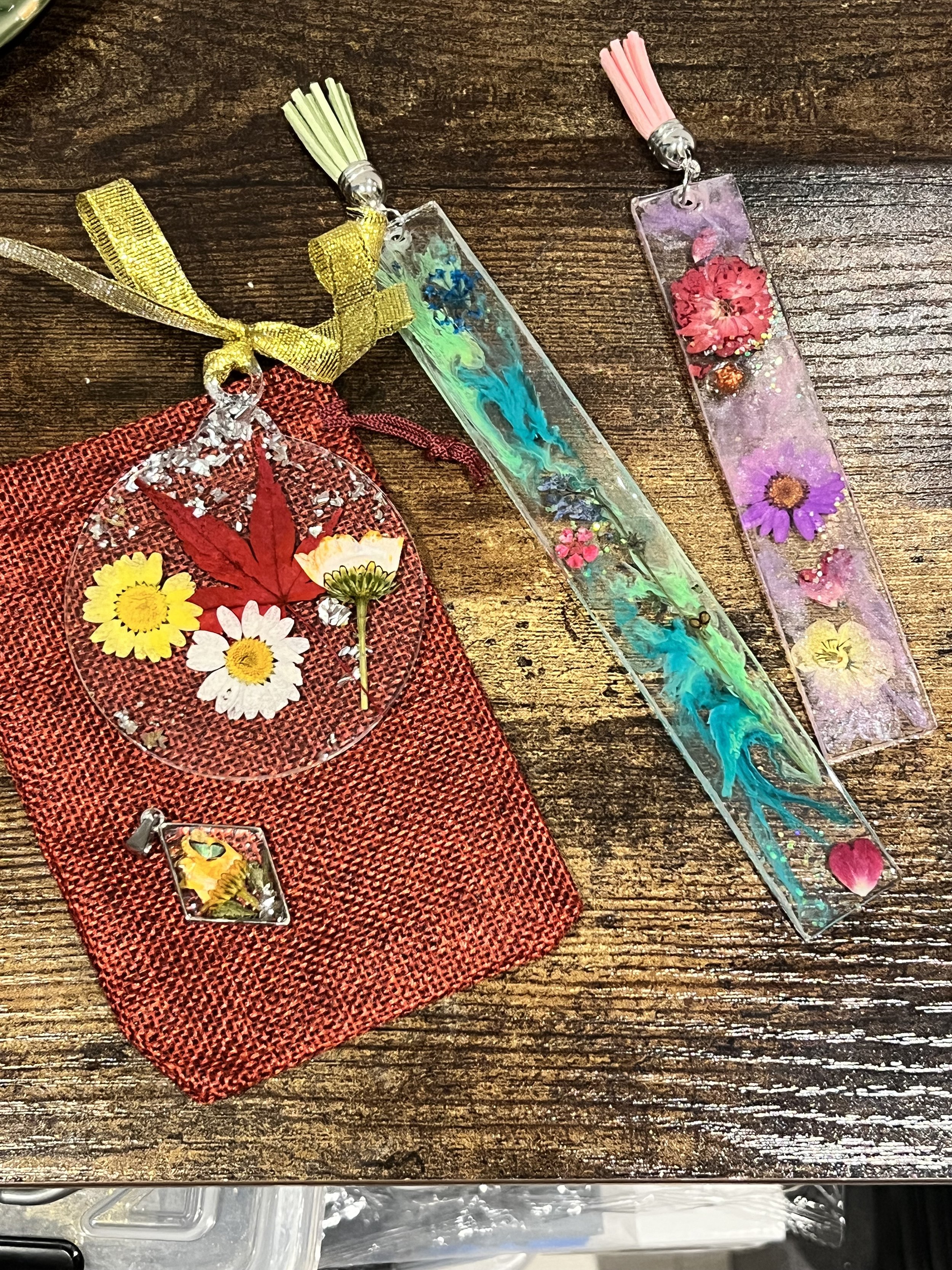 Decorative bookmarks and a jewelry charm with pressed flowers displayed on a wooden surface, with red textured fabric accessory.