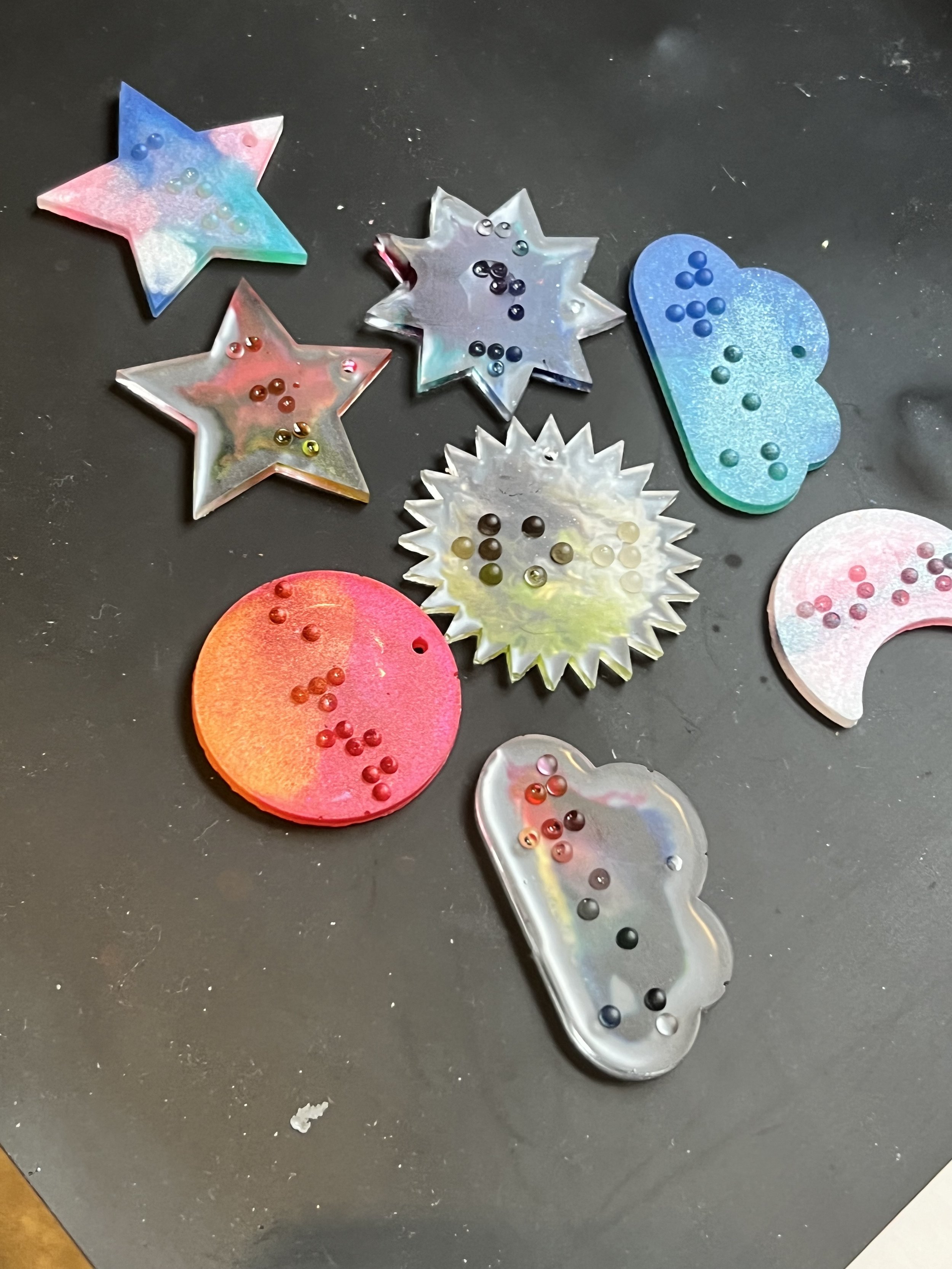 Colorful iridescent slime shapes including stars, clouds, a sun, and a moon, decorated with small colorful beads on a black surface.