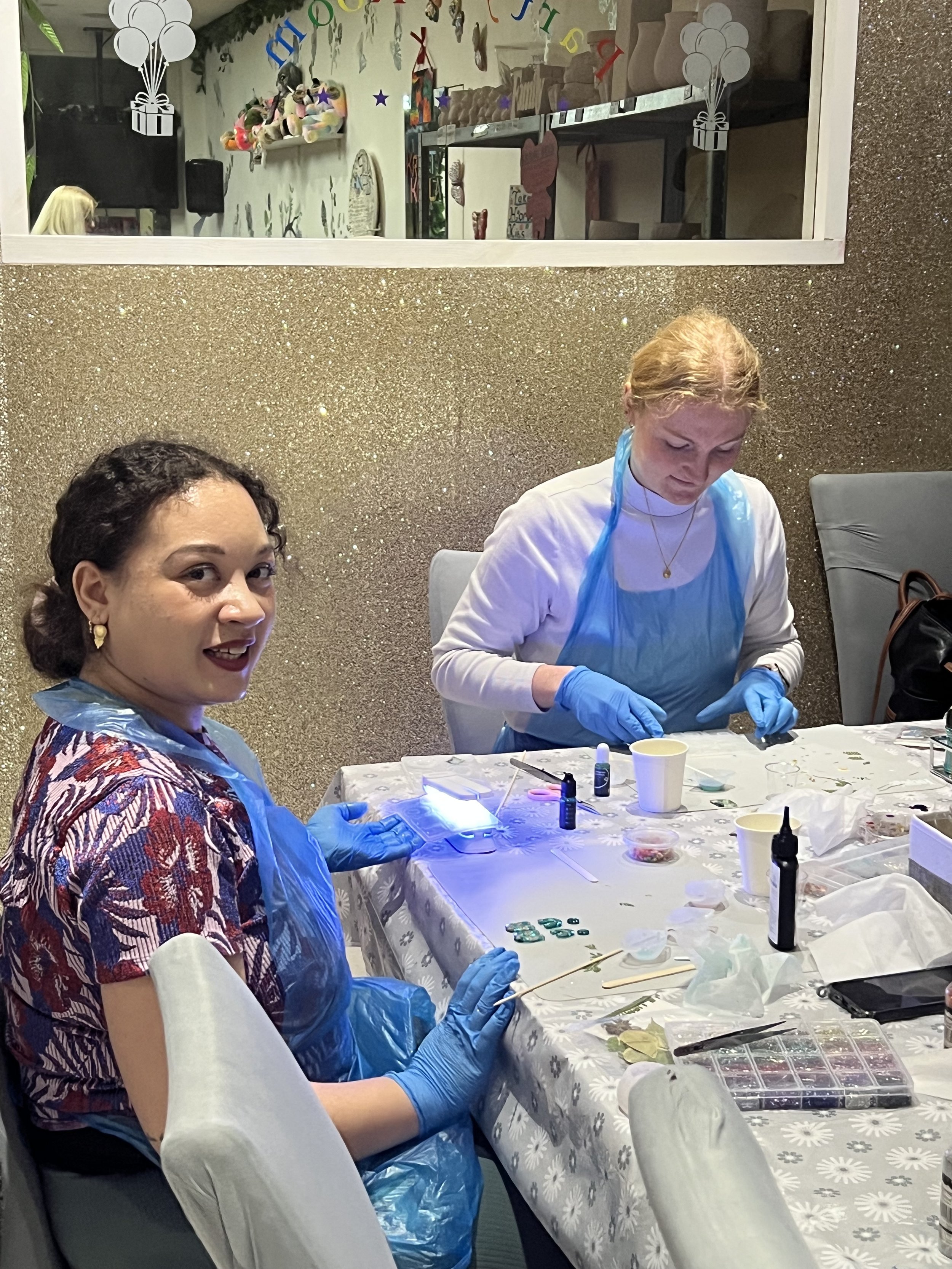 Two women seated at a table involved in a craft activity, with one woman smiling at the camera and the other focused, wearing gloves, with craft supplies on the table.
