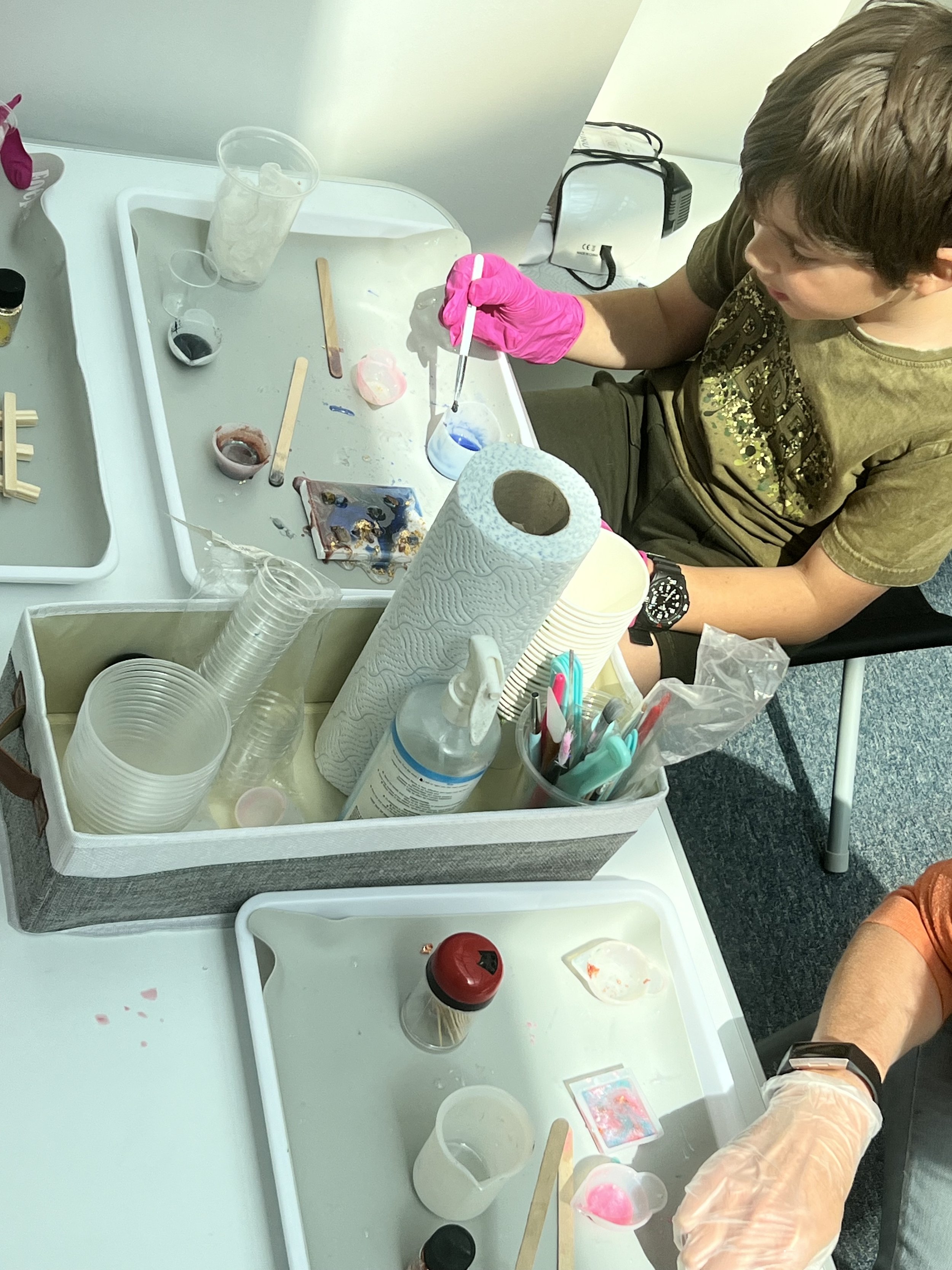 Person wearing pink gloves conducting a science experiment with colored liquids in test tubes, lab equipment on the table, and some cups and paper towels nearby.