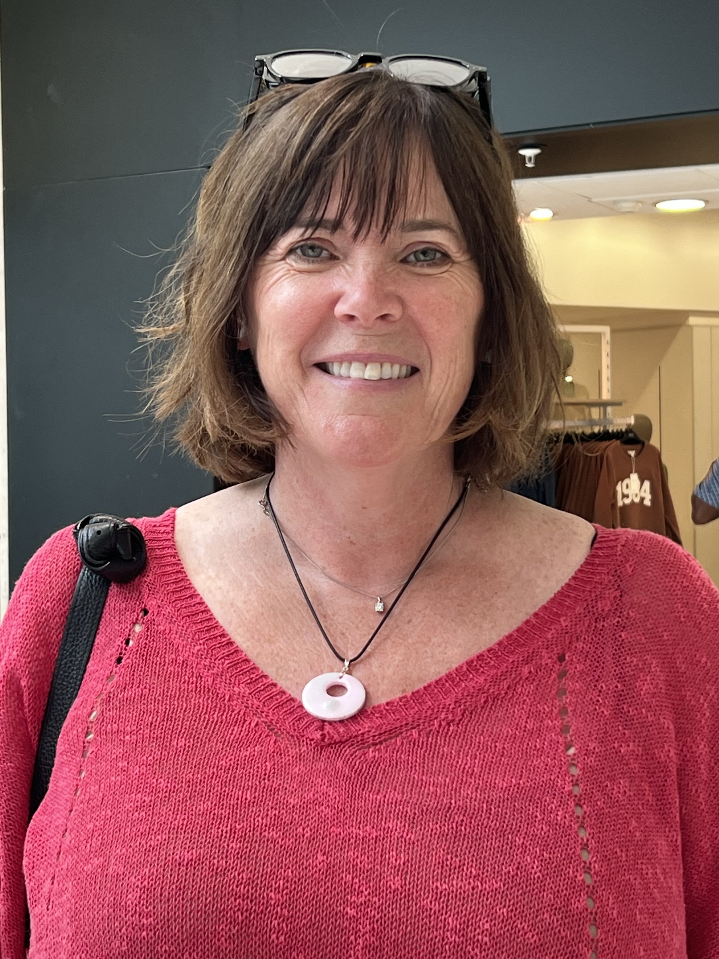 A woman with shoulder-length brown hair, wearing glasses on top of her head, a pink knitted sweater, and two necklaces, smiling in an indoor setting.
