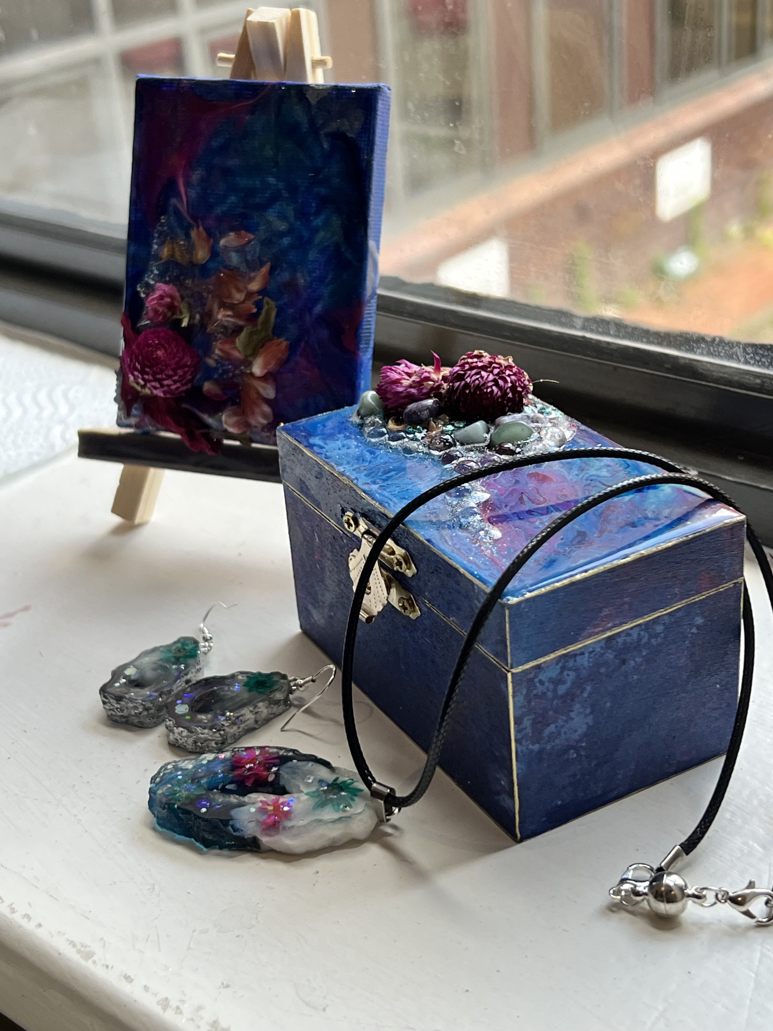 Decorative jewelry display with a blue box, a necklace with a floral pendant, earrings, and a small easel with an abstract painting, all arranged on a windowsill.