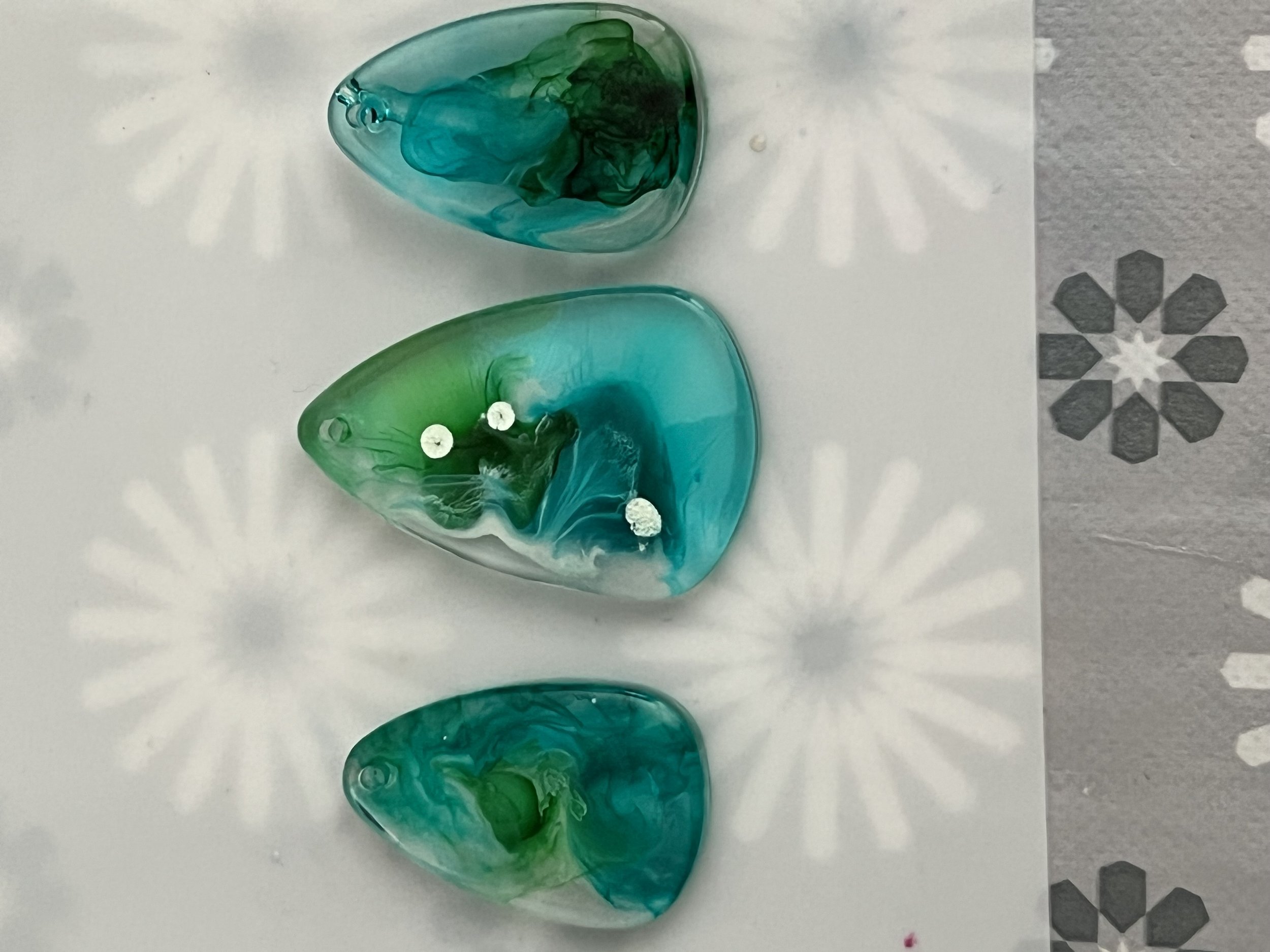 Three glass heart-shaped objects in green and blue shades, with the middle one decorated with white dots, placed on a white surface with a shadow pattern underneath.