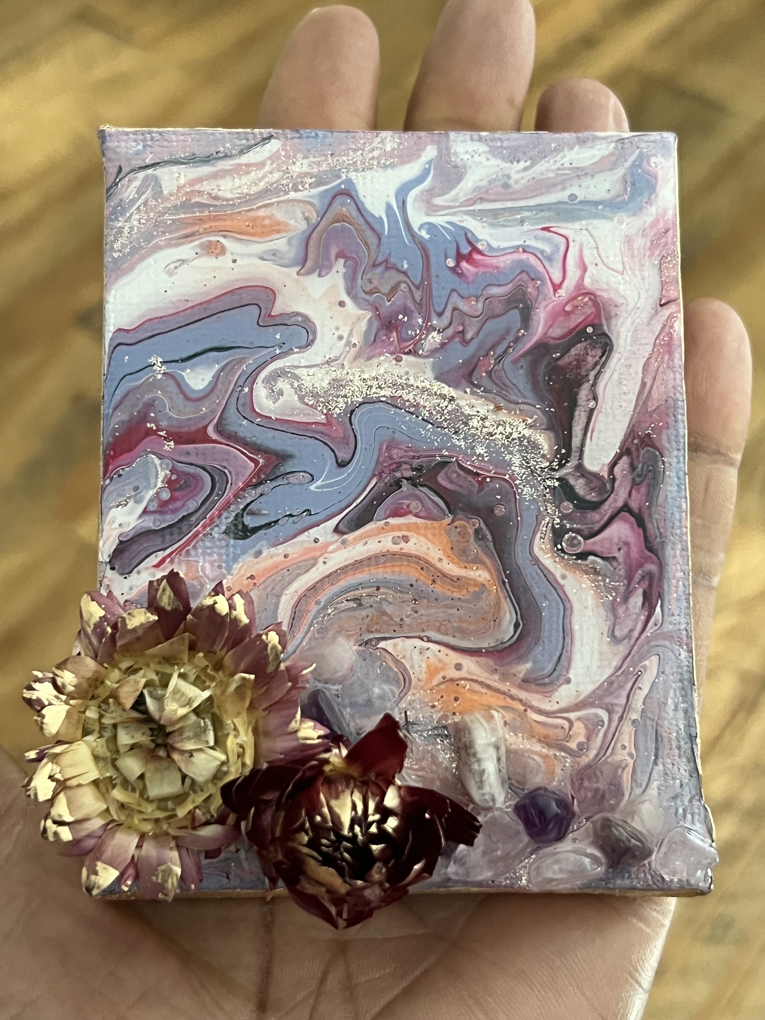 A person's hand holding a small canvas with an abstract, marbled painting in shades of pink, purple, blue, black, and white, decorated with dried flowers at the bottom left corner.