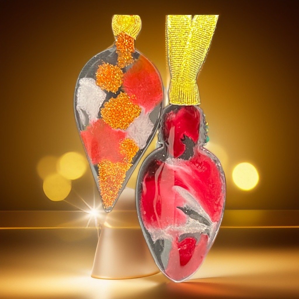Artistic glass sculptures of a spoon and a heart, filled with red, gold, silver, and white colorful materials, illuminated against a golden background with bokeh lights.
