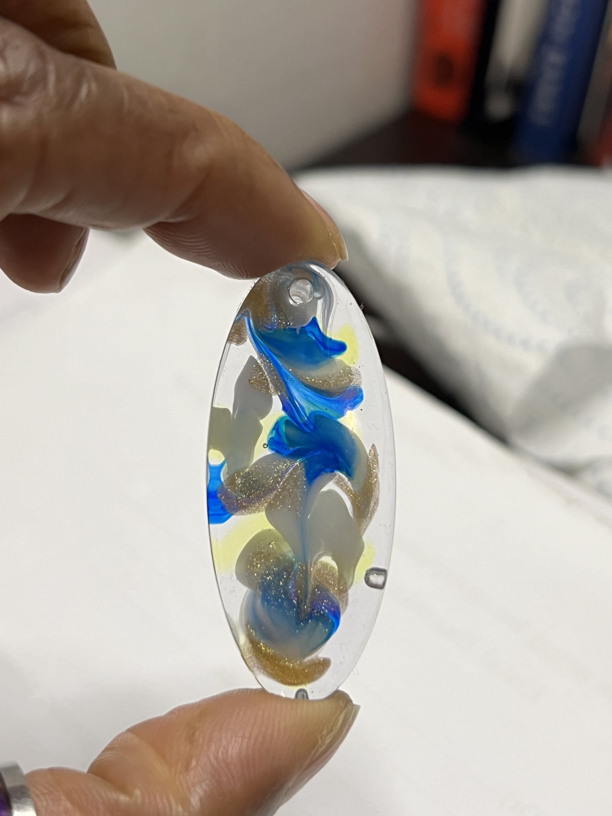 Hand holding an oval-shaped glass ornament with swirls of blue, white, yellow, and gold glitter inside.
