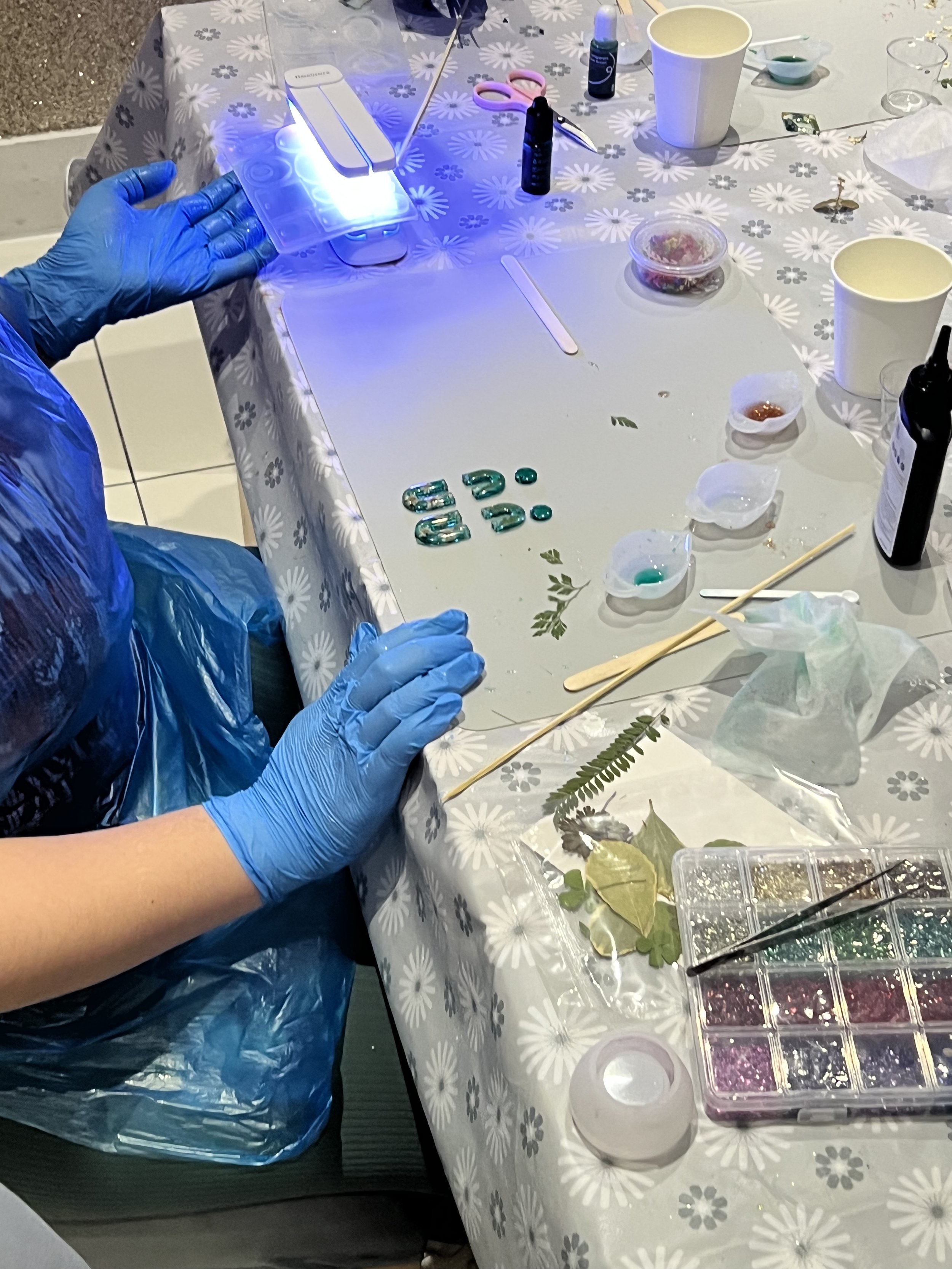 A person wearing blue gloves and a plastic apron working at a table with craft supplies, including paint, brushes, glitter, leaves, and decorative items, possibly creating a decorated glass or ornament.