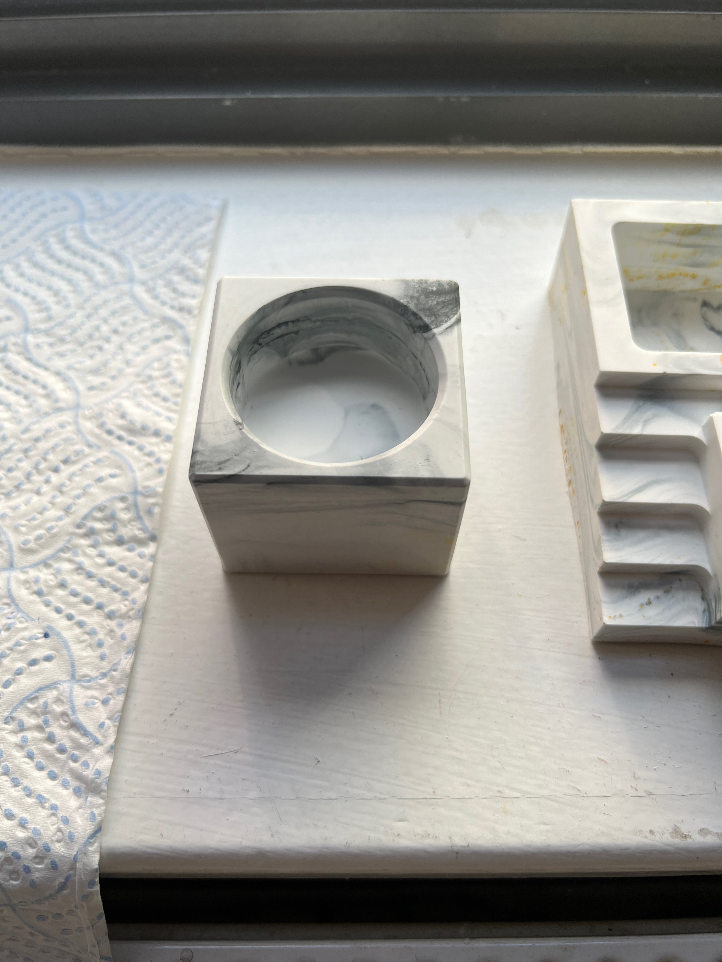 Marble cube with a hollowed-out top, placed on a white surface near a paper towel and another white marble object with rectangular and curved shapes.
