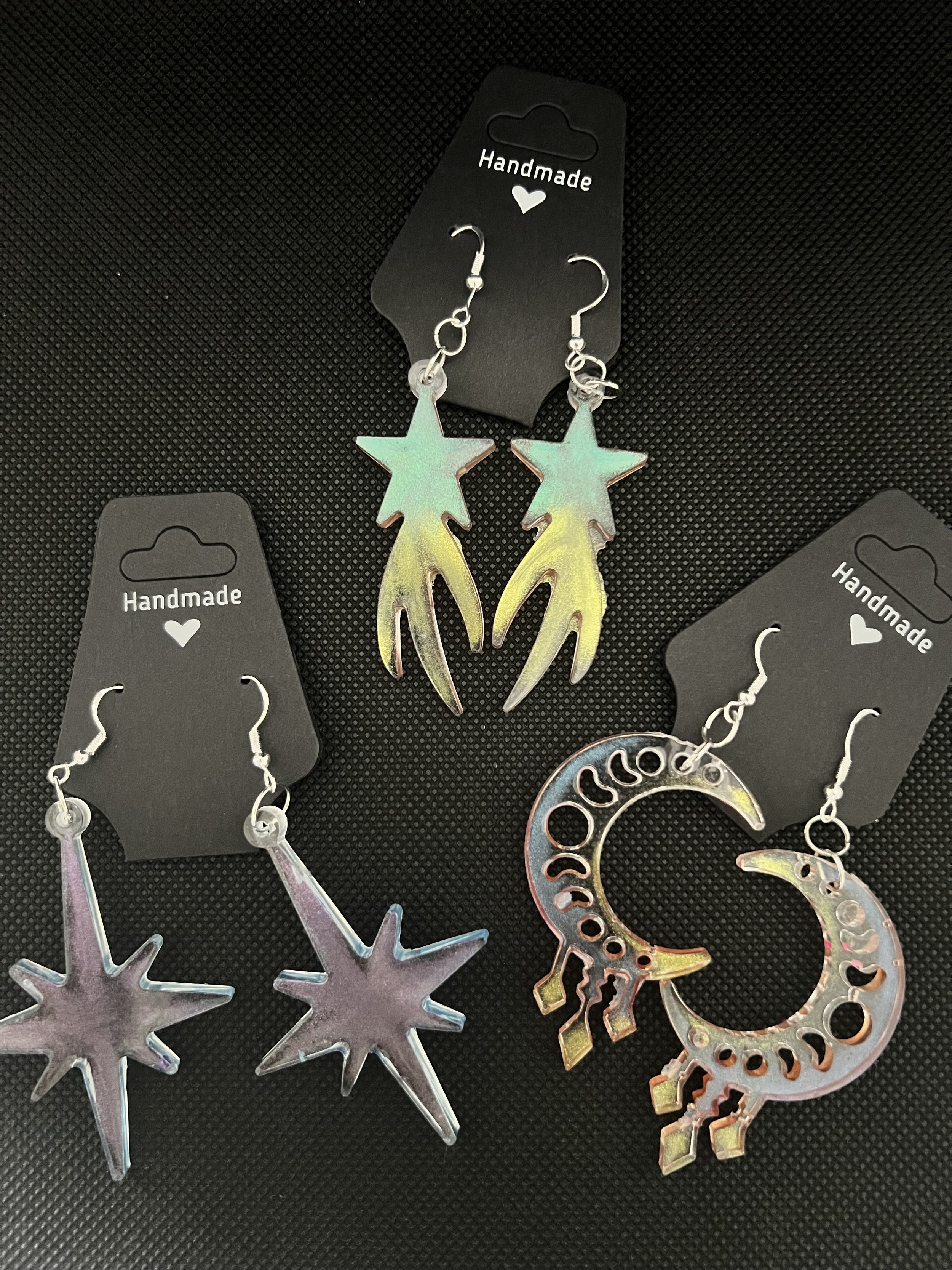 Three pairs of colorful handmade earrings on black display cards, including star, starburst, and crescent moon designs.