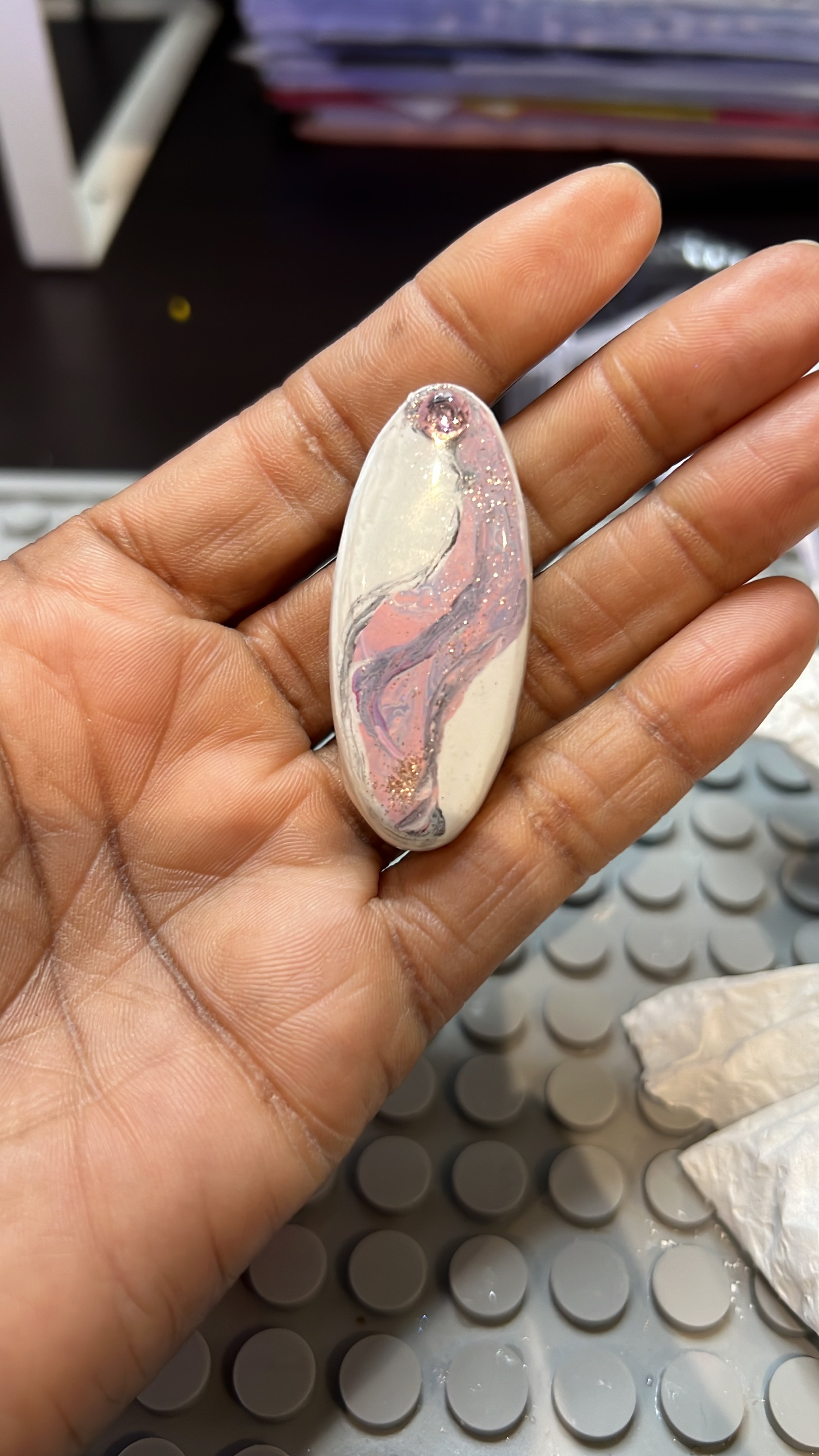 Hand holding an oval-shaped, marble-patterned resin or stone object with pink, purple, and white swirls and glitter accents.