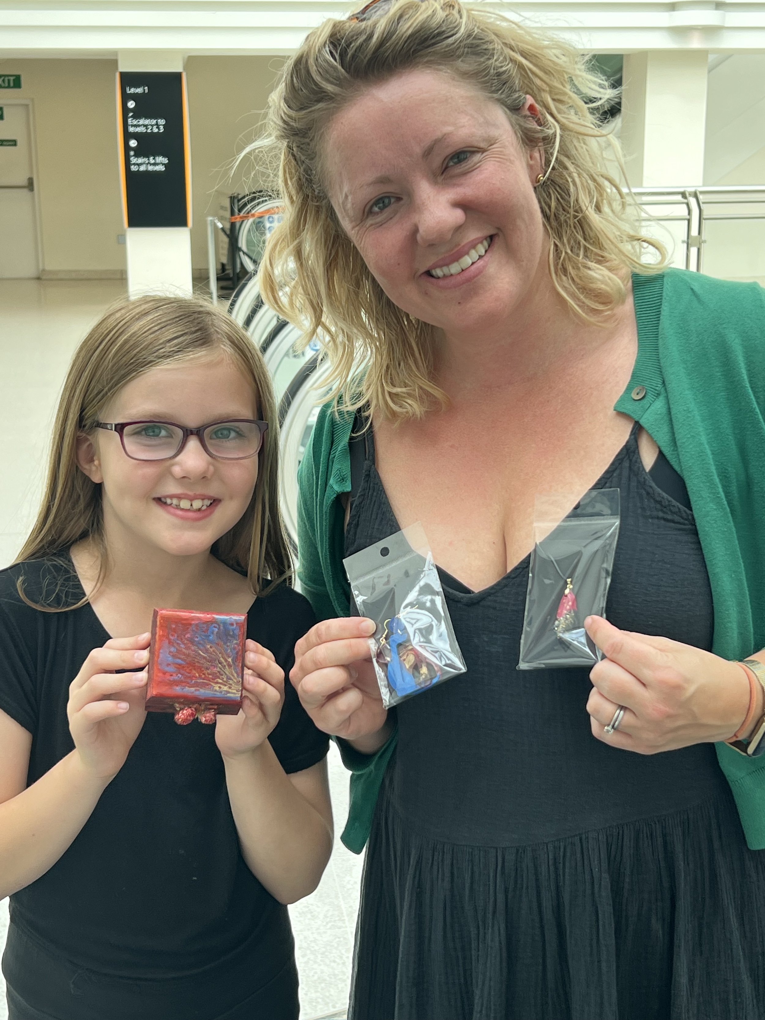 A woman and a girl standing together in an indoor setting, both smiling and holding small packaged items.