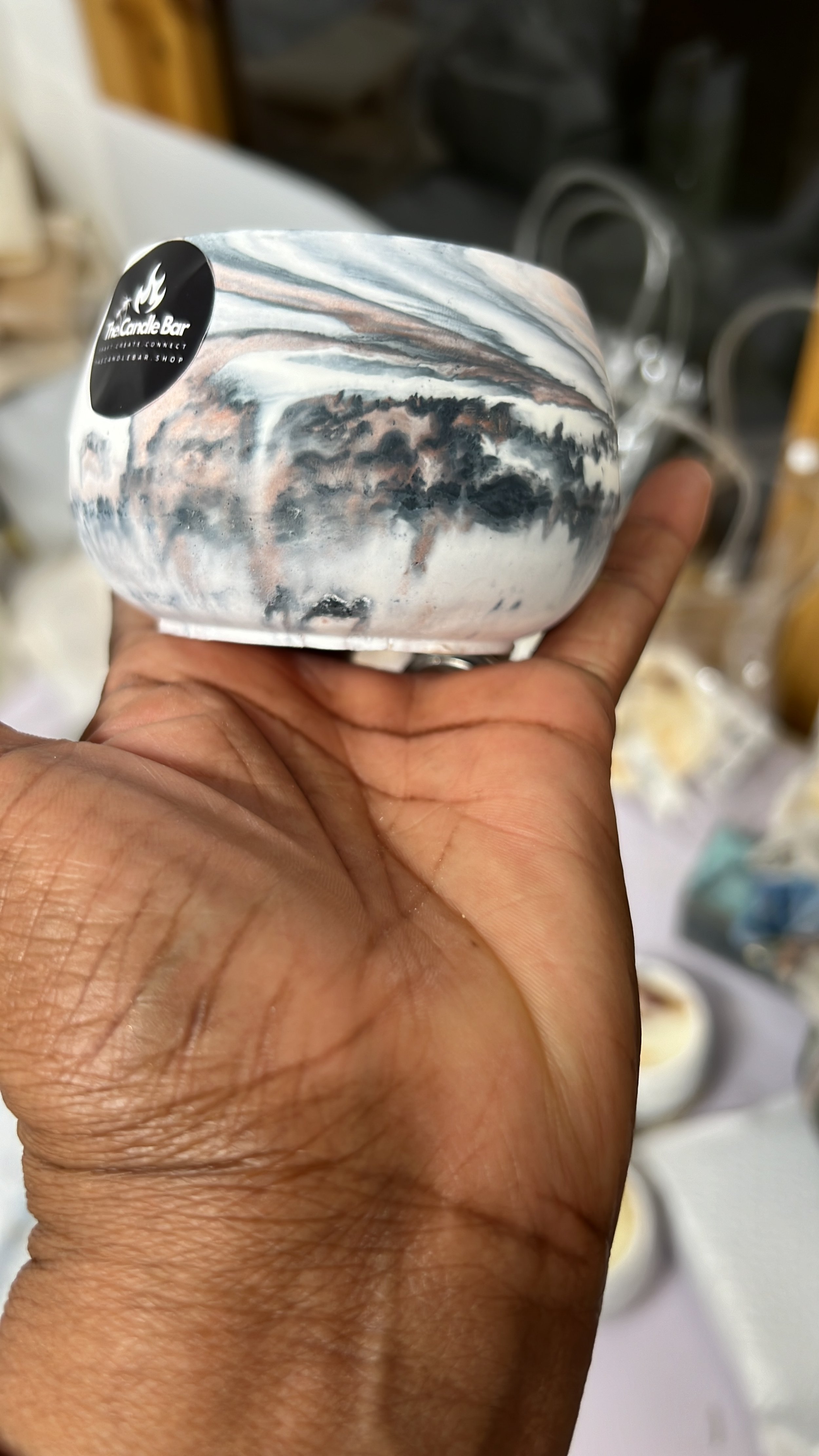 Hand holding a marble-colored candle in a small bowl-shaped container with a black sticker that reads "The Candle Bar".