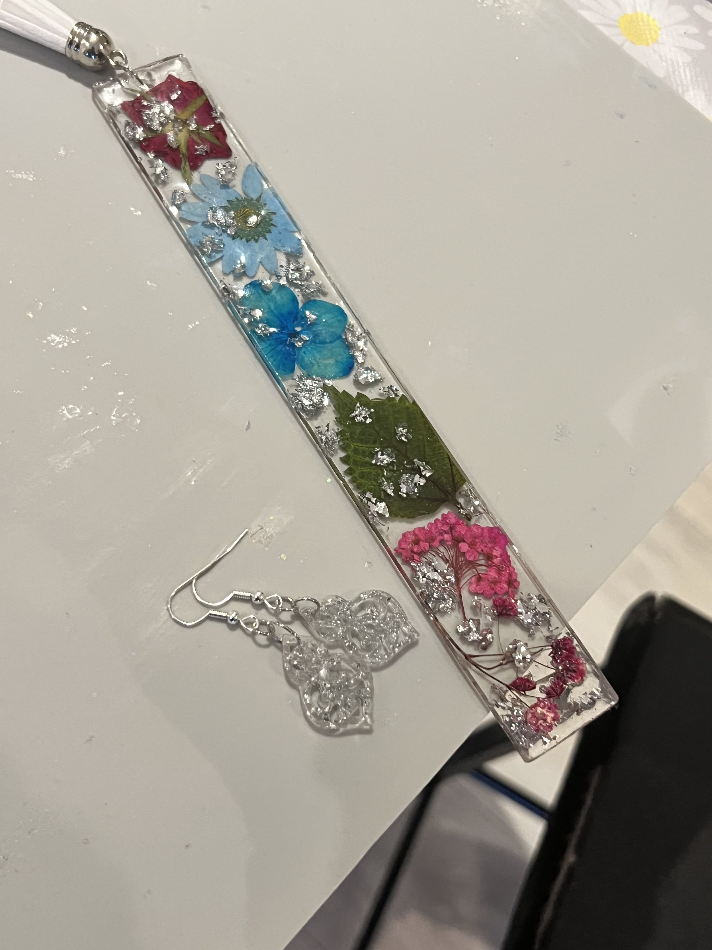 A clear rectangular resin bracelet with embedded colorful dried flowers and glitter, accompanied by matching clear butterfly earrings, on a white surface.
