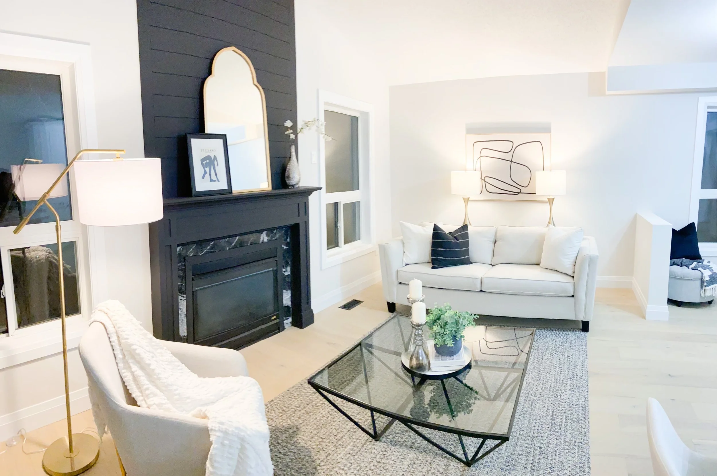 Living room with a white sofa, black and white pillows, a glass coffee table with candles and a plant, a fireplace with a black mantel and a decorative mirror, a white armchair with a blanket, and lamps on side tables, with light-colored walls and ha