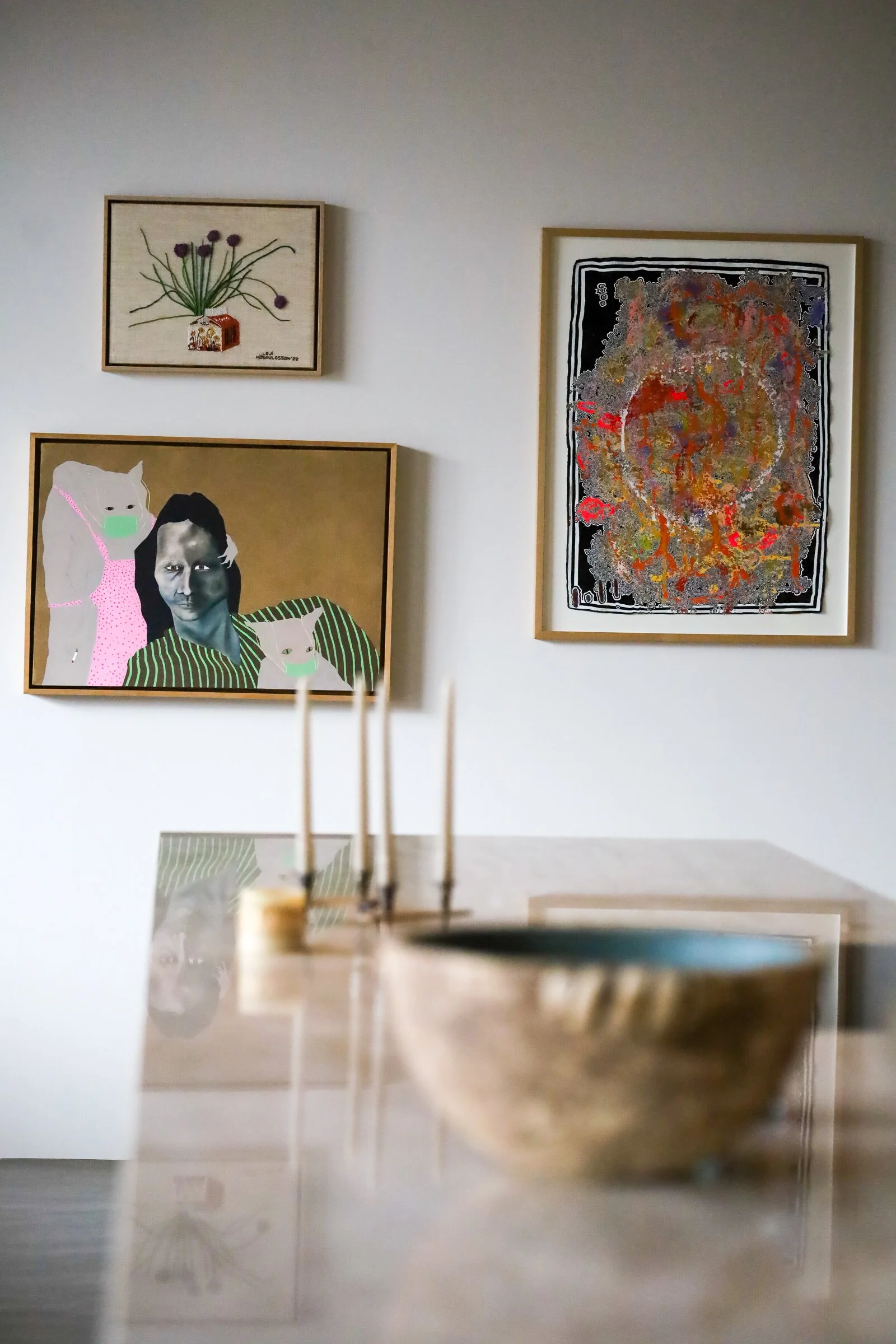 Abstract art gallery interior with three paintings on a wall; one depicts a plant, another a figure with a cat, and the third is a colorful abstract design. In the foreground, a bowl and incense sticks are on a reflective surface.