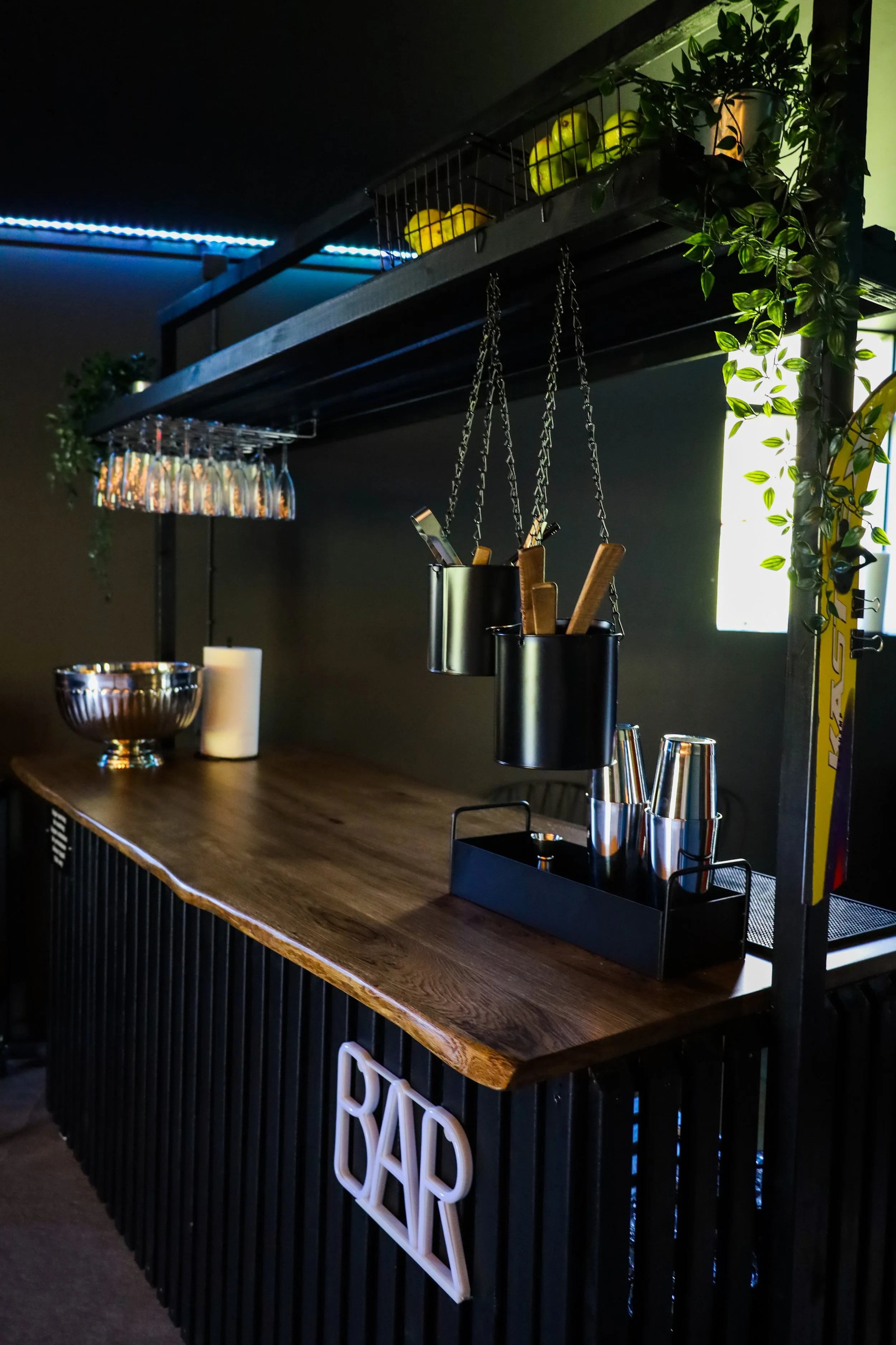 A modern bar setup with a wooden countertop, hanging metal containers, and bar tools. Glasses are hung upside-down under the frame, and there are decorative elements like a metal bowl, paper towel, and potted plants. The word 'BAR' is displayed on the front.