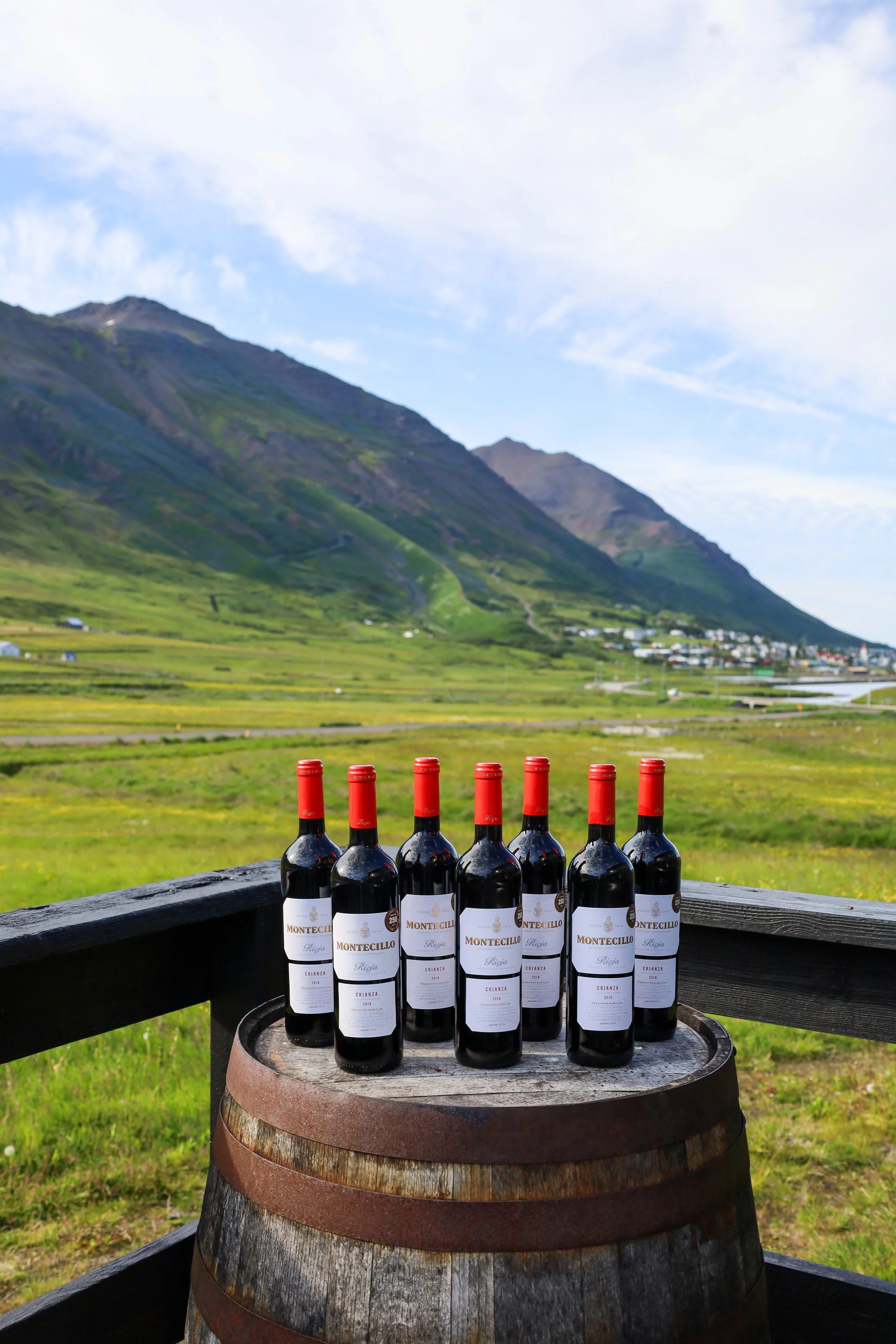 Nine bottles of wine with red caps on a wooden barrel, set against a grassy mountain landscape.