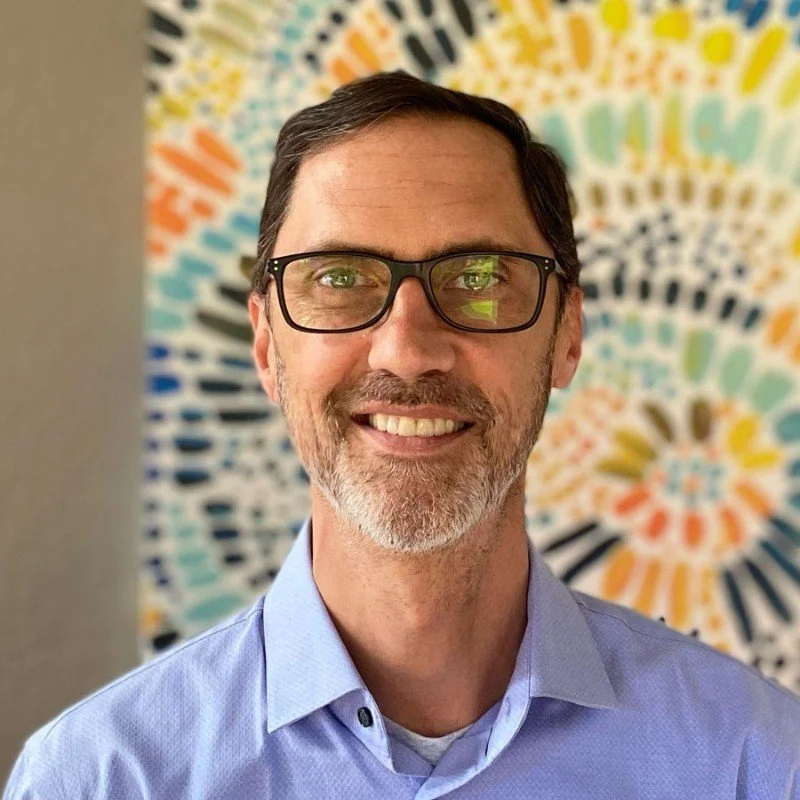 A man with dark hair and glasses, smiling, wearing a light blue button-up shirt. The background features a vibrant, colorful, abstract pattern.