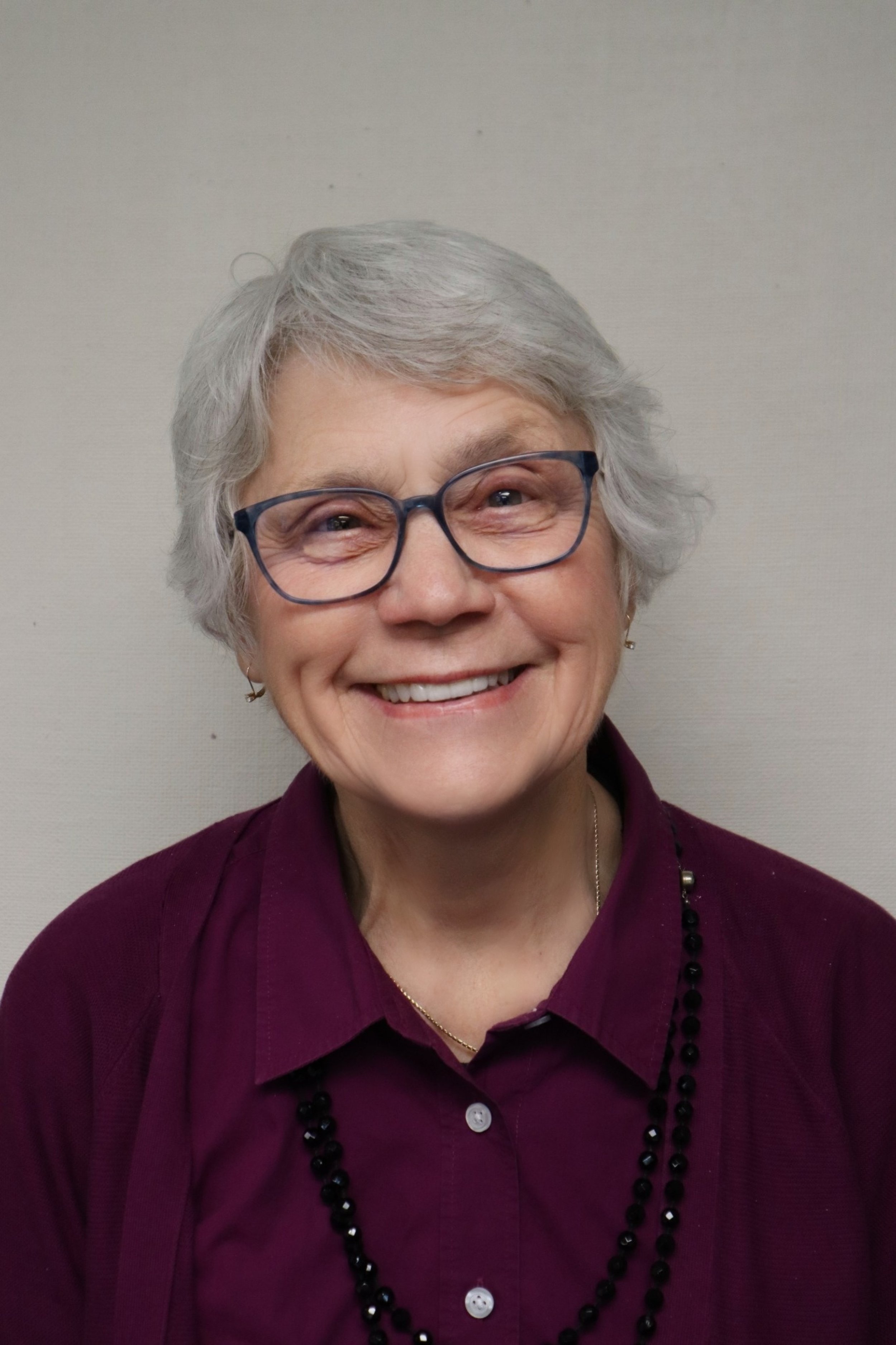 Portrait of an elderly woman with short gray hair, glasses, and a warm smile, wearing a burgundy blouse and black beaded necklace, standing against a plain light-colored wall.