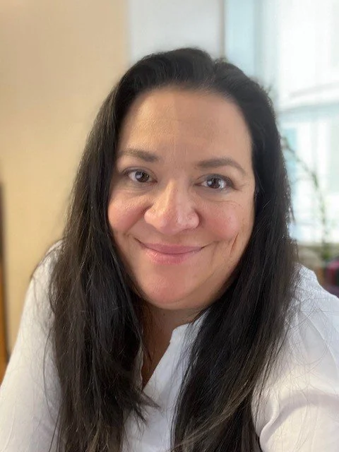 Close-up of a smiling woman with long dark hair wearing a white top, indoors with a blurred background and natural light.