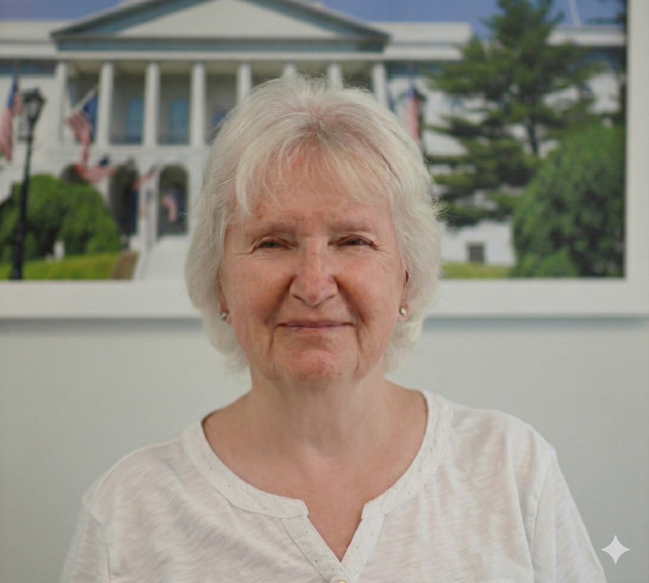 An elderly woman with white hair, wearing a white shirt, smiling softly in front of a large white house with multiple American flags.