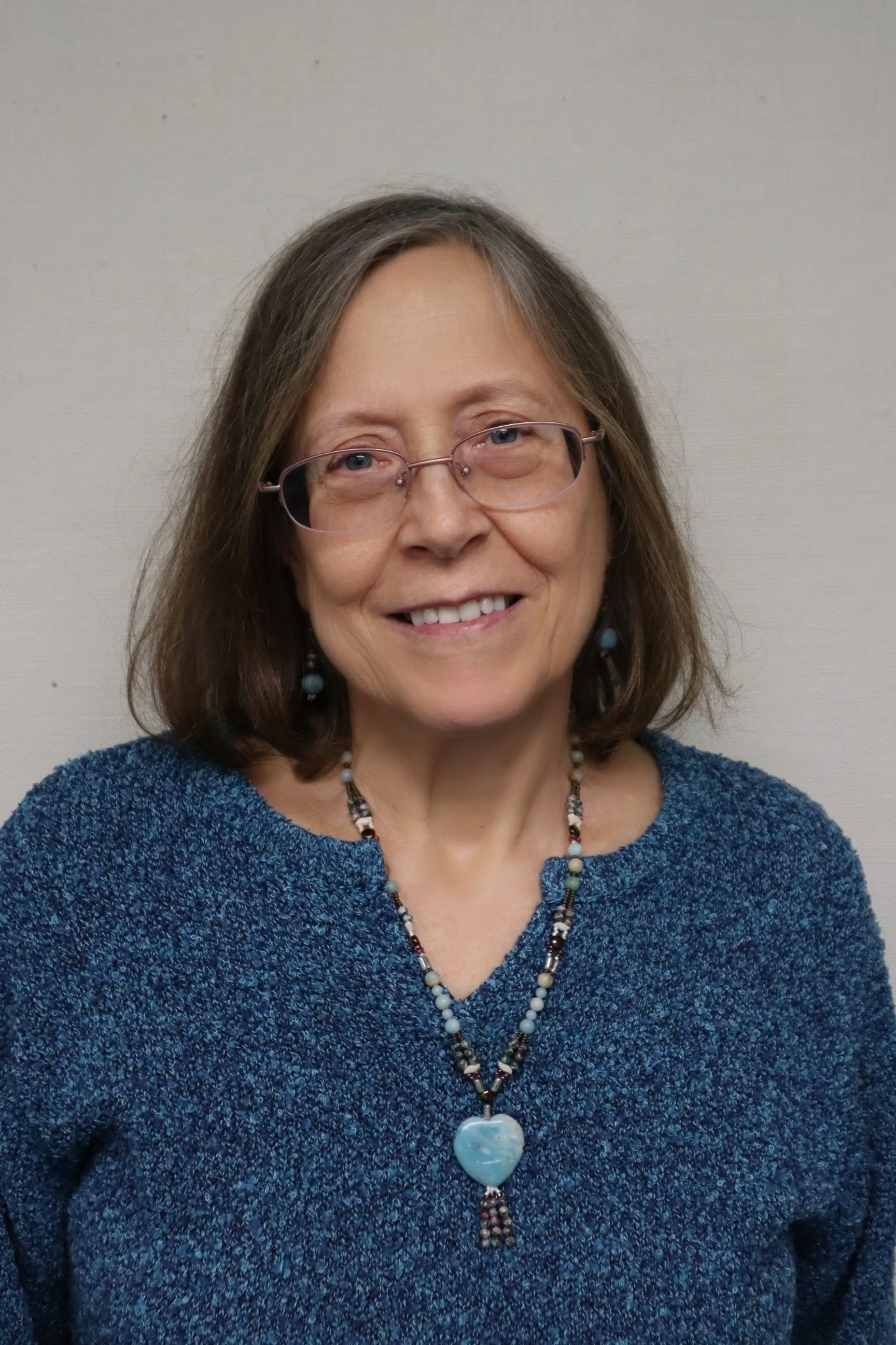 A smiling middle-aged woman with glasses, shoulder-length brown hair, wearing a blue sweater, beaded necklace with a heart-shaped pendant, and matching earrings, standing against a plain light-colored wall.