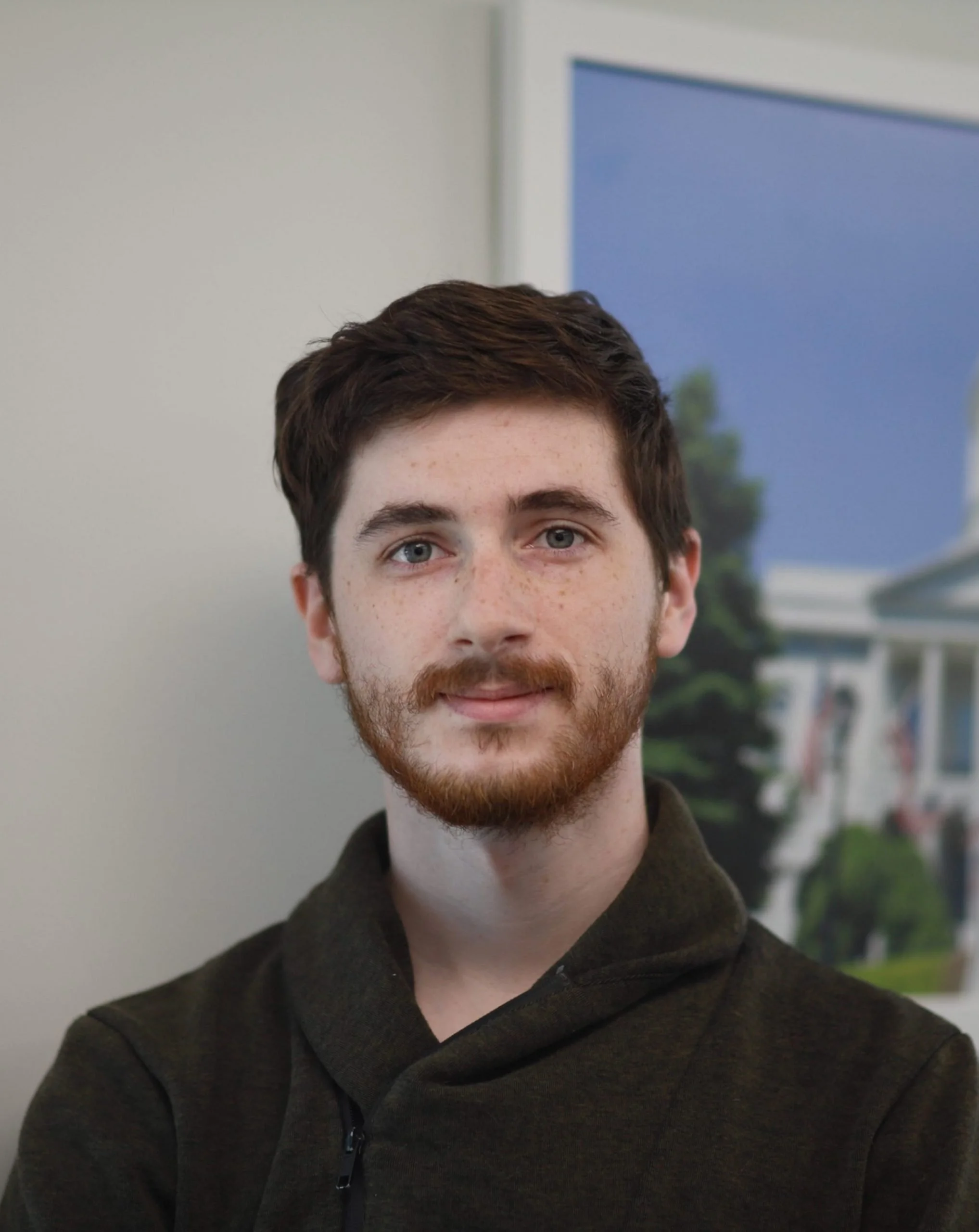 Portrait of a young man with brown hair and a beard, wearing a dark zip-up hoodie, standing in front of a blurred background with a photo of a house and trees.