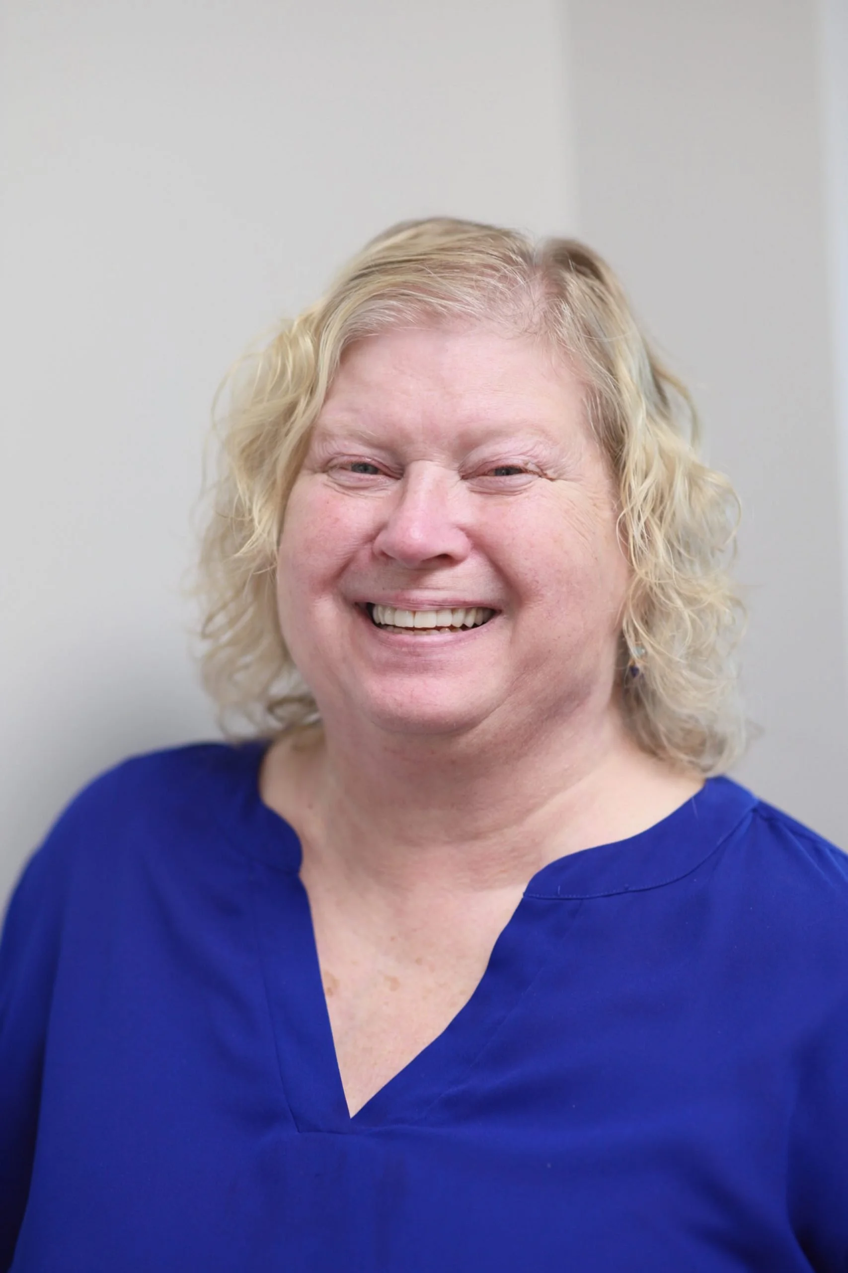 A smiling woman with blonde, curly hair wearing a blue top standing against a plain light-colored wall.