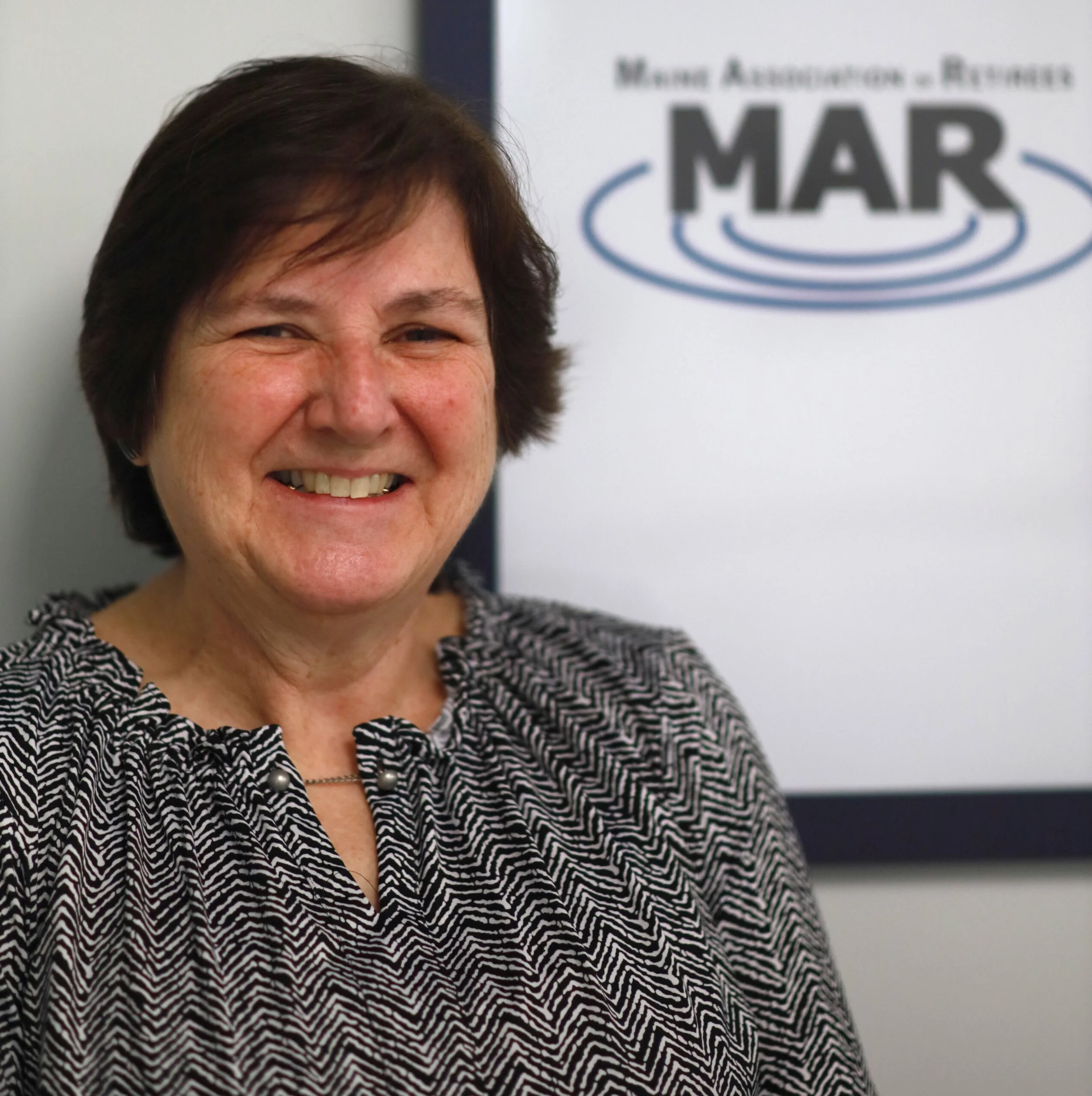 Smiling woman with short brown hair wearing a black and white patterned blouse, standing in front of a sign with the logo MAR and the words 'Mare Associates & Engineering'.
