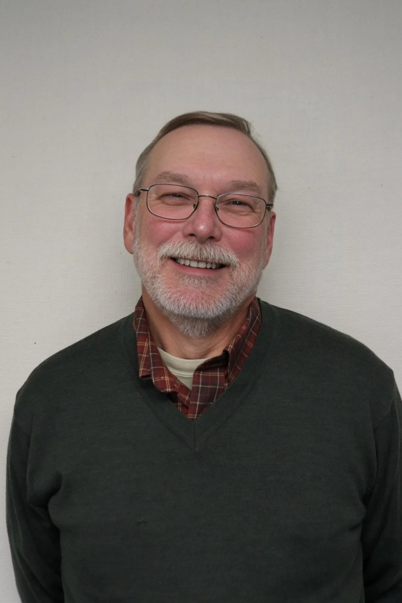 A smiling middle-aged man with glasses, a beard, and gray hair, wearing a dark sweater over a plaid shirt, standing against a plain light-colored wall.