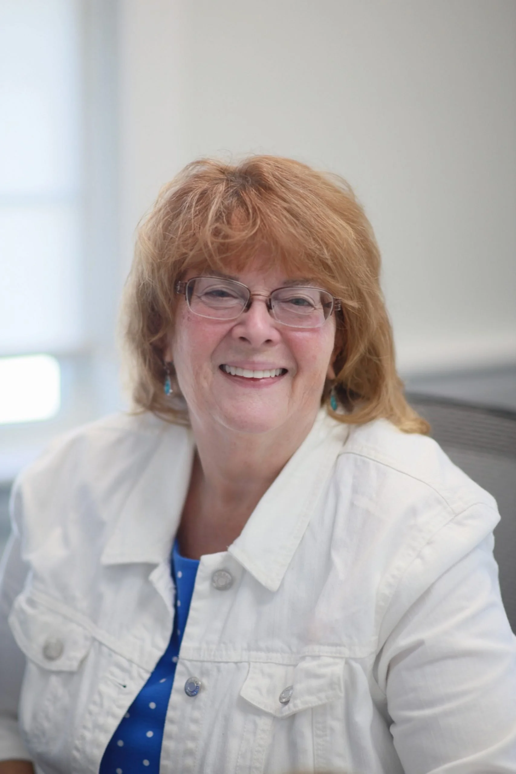 A woman with curly red hair and glasses smiling at the camera, dressed in a white jacket over a blue top, in an indoor setting.