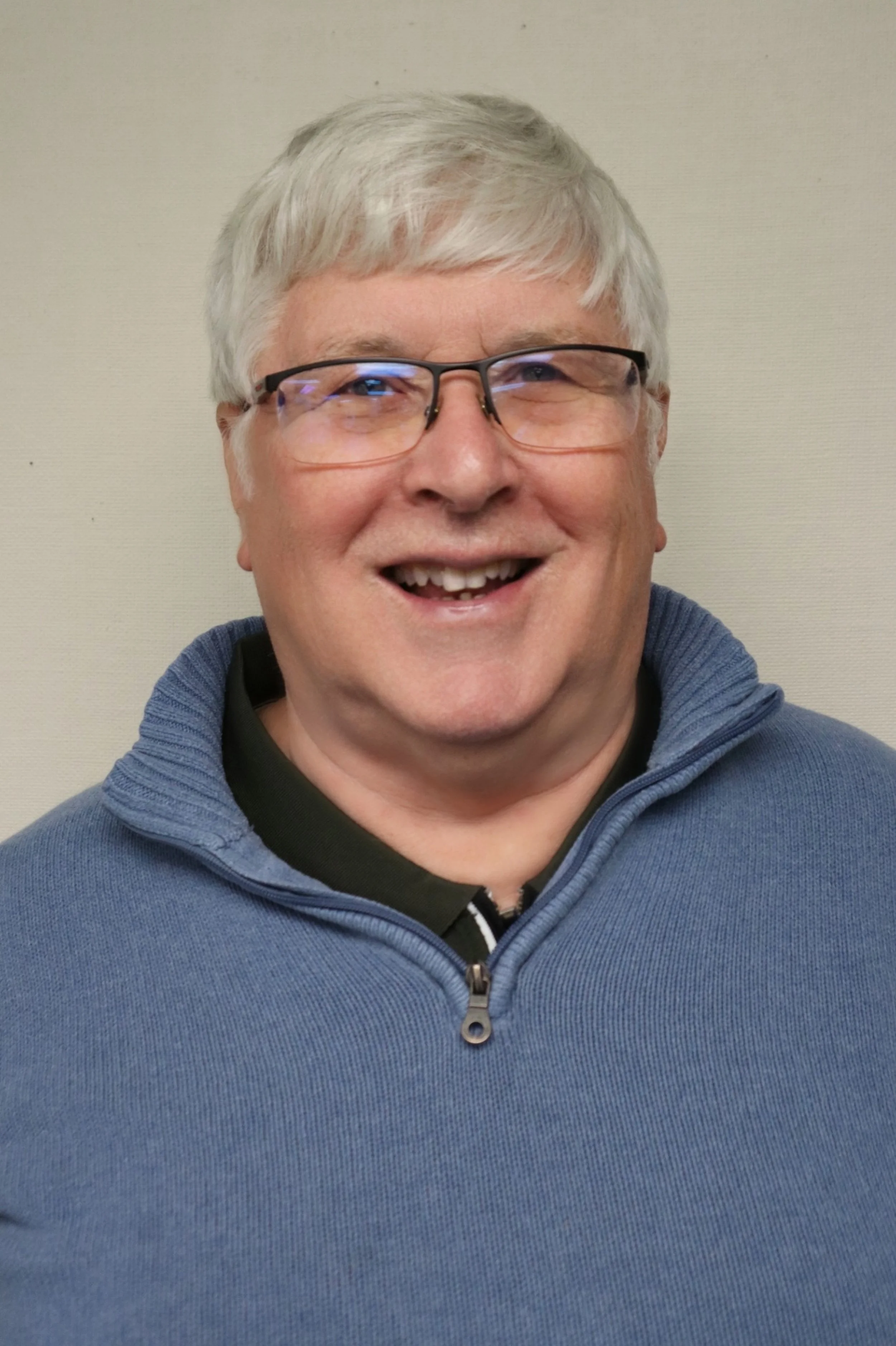 A smiling man with gray hair, glasses, wearing a blue zip-up sweater over a black collared shirt, standing against a plain light-colored wall.