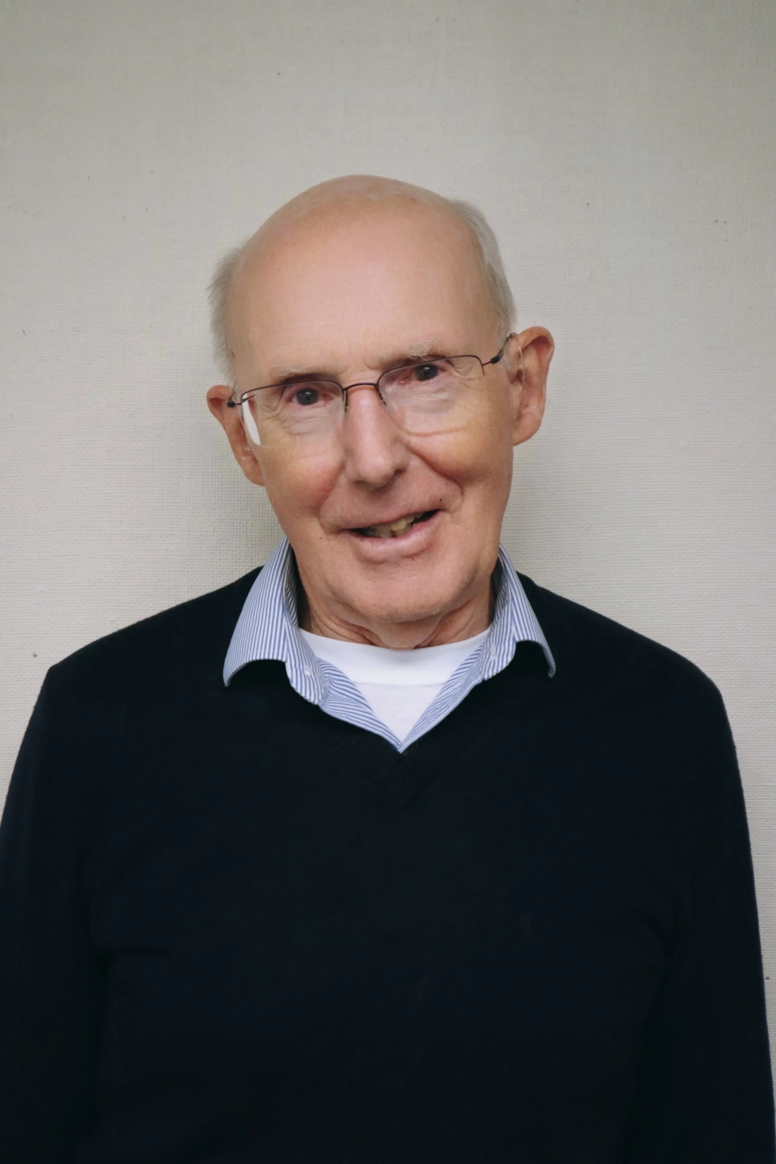 An elderly man with glasses, wearing a collared shirt and black sweater, standing against a plain beige background.