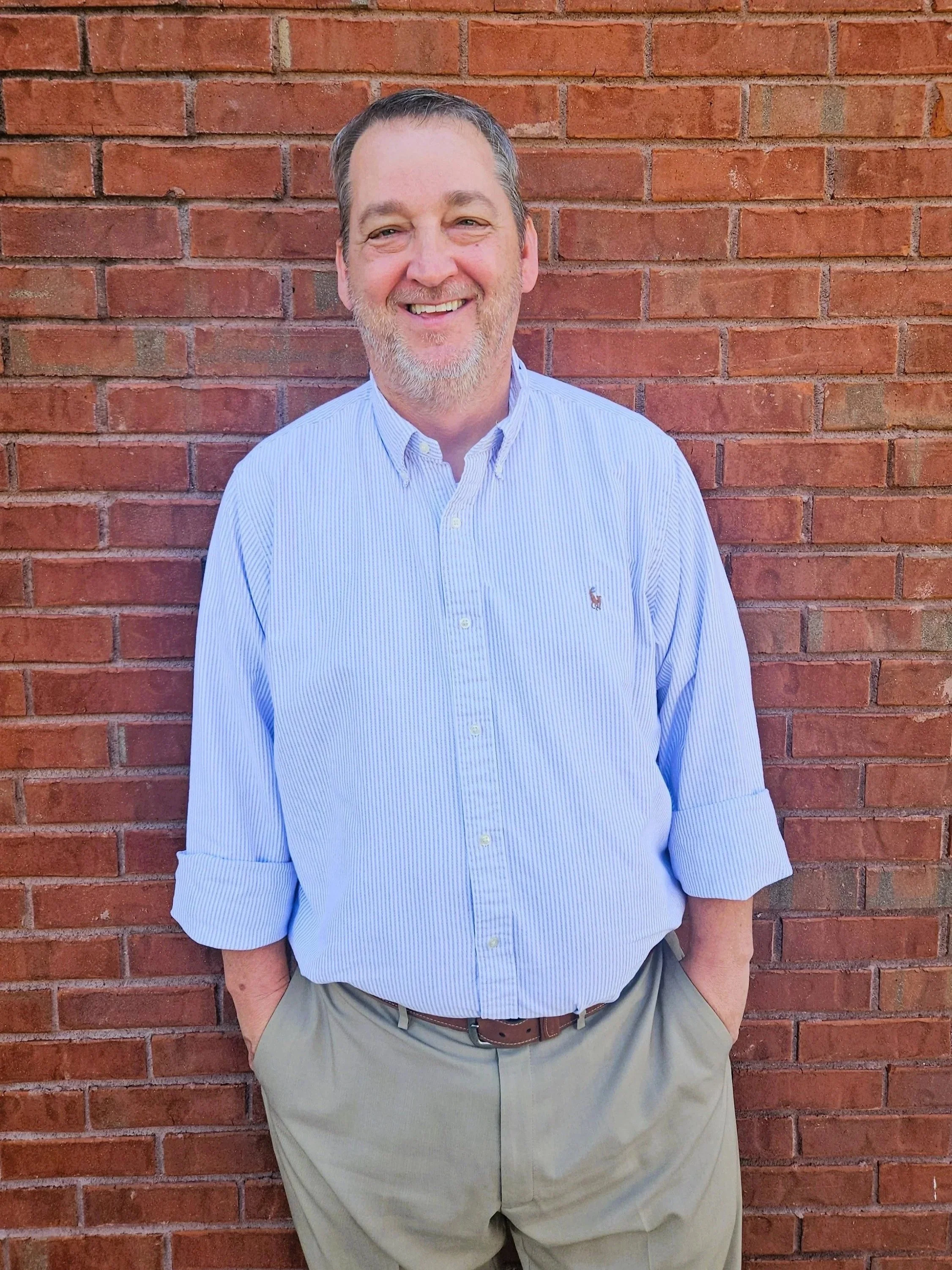 A smiling man with gray hair and a beard, dressed in a light blue striped button-up shirt with rolled-up sleeves and khaki pants, standing in front of a brick wall.