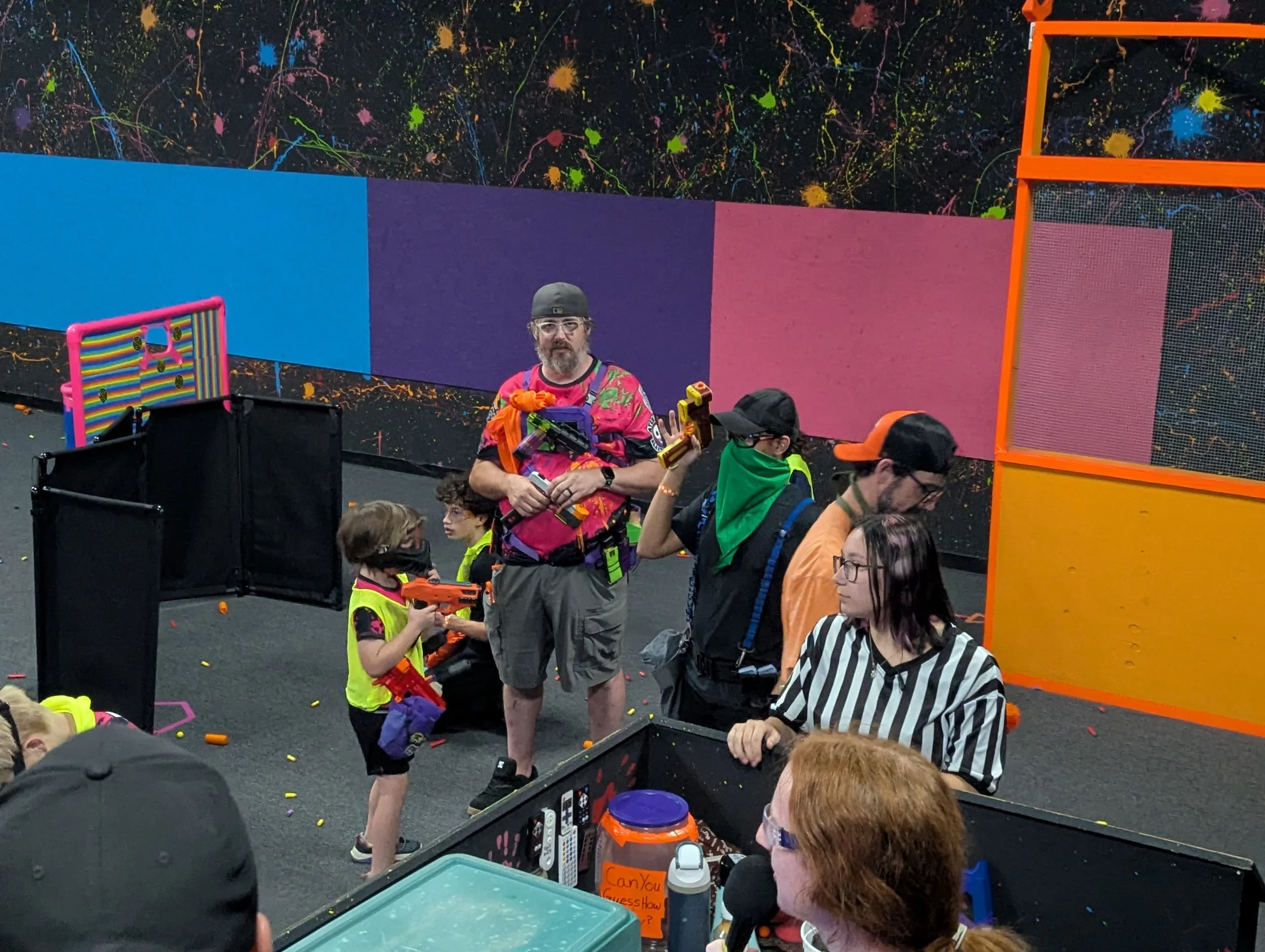 A group of children and adults with toy guns and colorful clothing inside an indoor Nerf arena with a black wall decorated with colorful paint splatters, orange netting, and foam barriers.