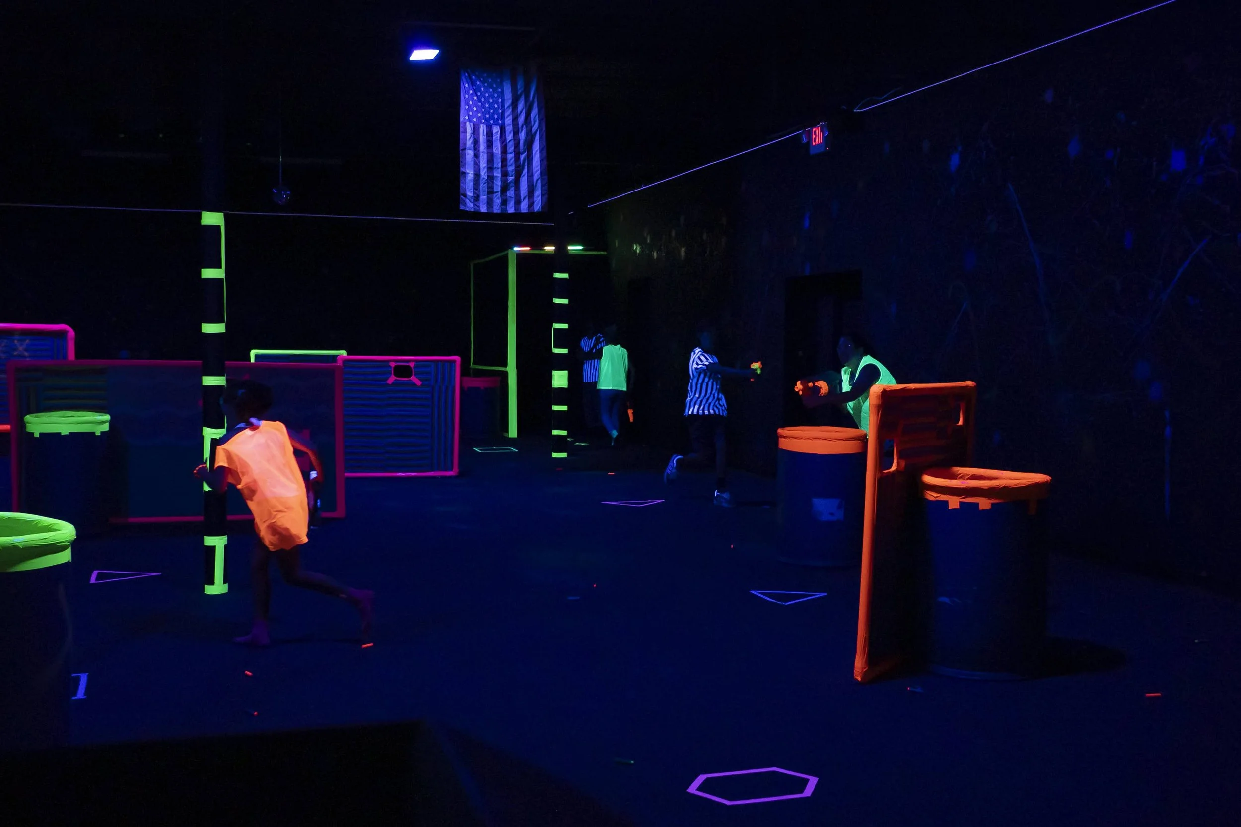 People playing laser tag in a dark, glow-in-the-dark arena with neon-lit obstacles and walls.