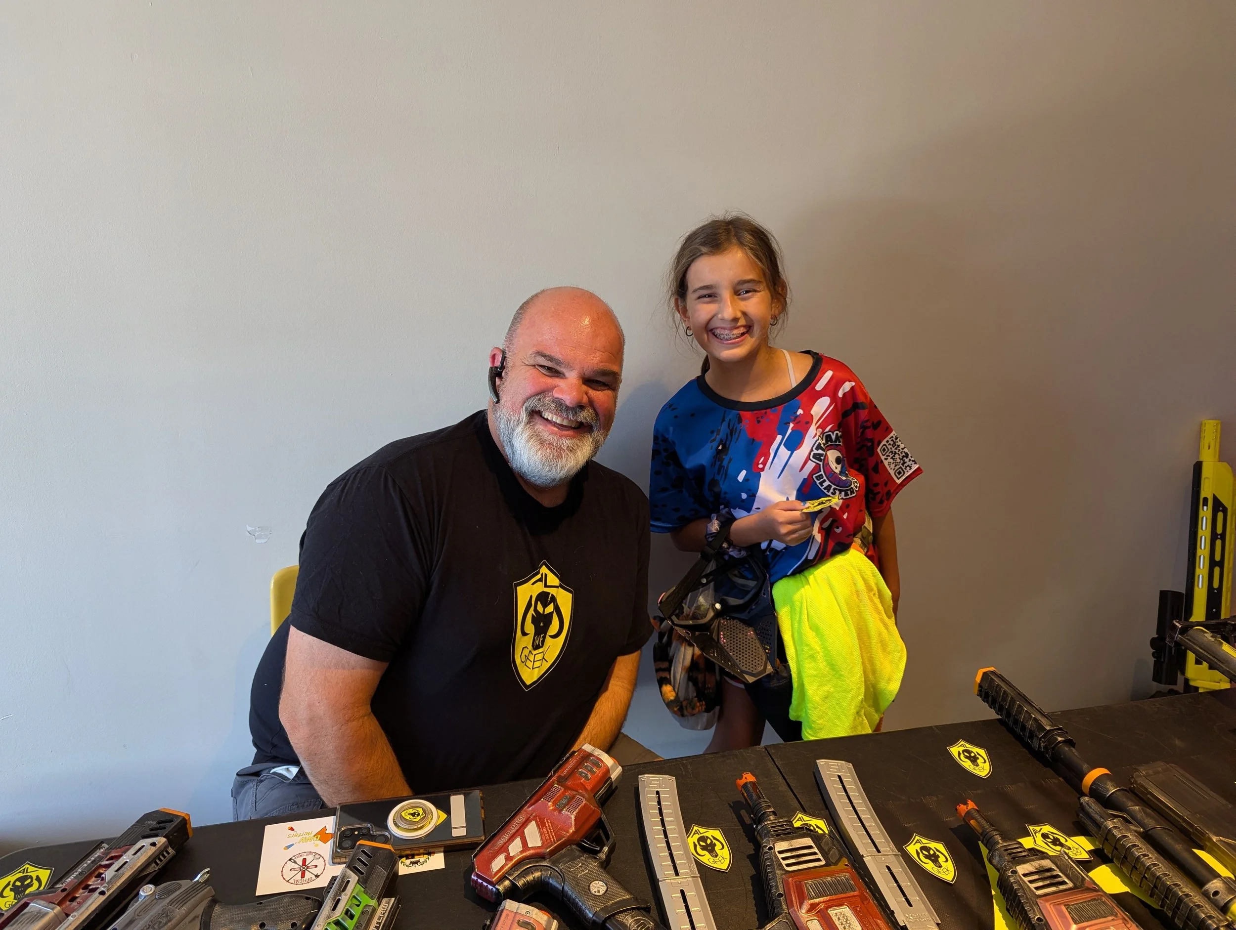 A man with a beard and a headset smiling next to a young girl with braces, both standing behind a table with various tools and equipment, including power drills and tool magazines, in a room with a plain wall.