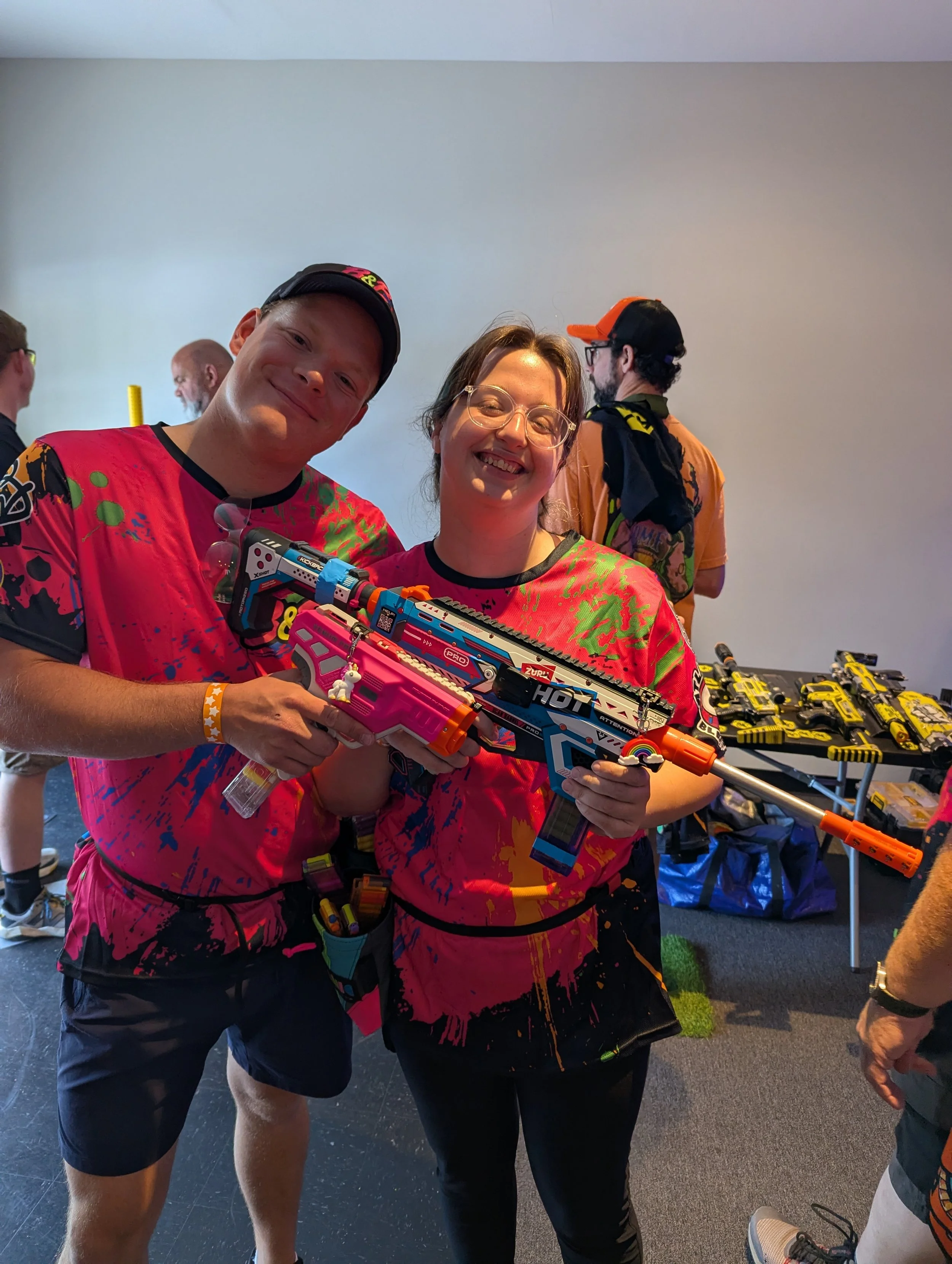 Two individuals smiling, holding a colorful Nerf blaster at a table with Nerf toys in a room with plain walls. One person is wearing glasses and a black and orange cap, and the other is wearing a colorful shirt and a black cap. Other people are in th