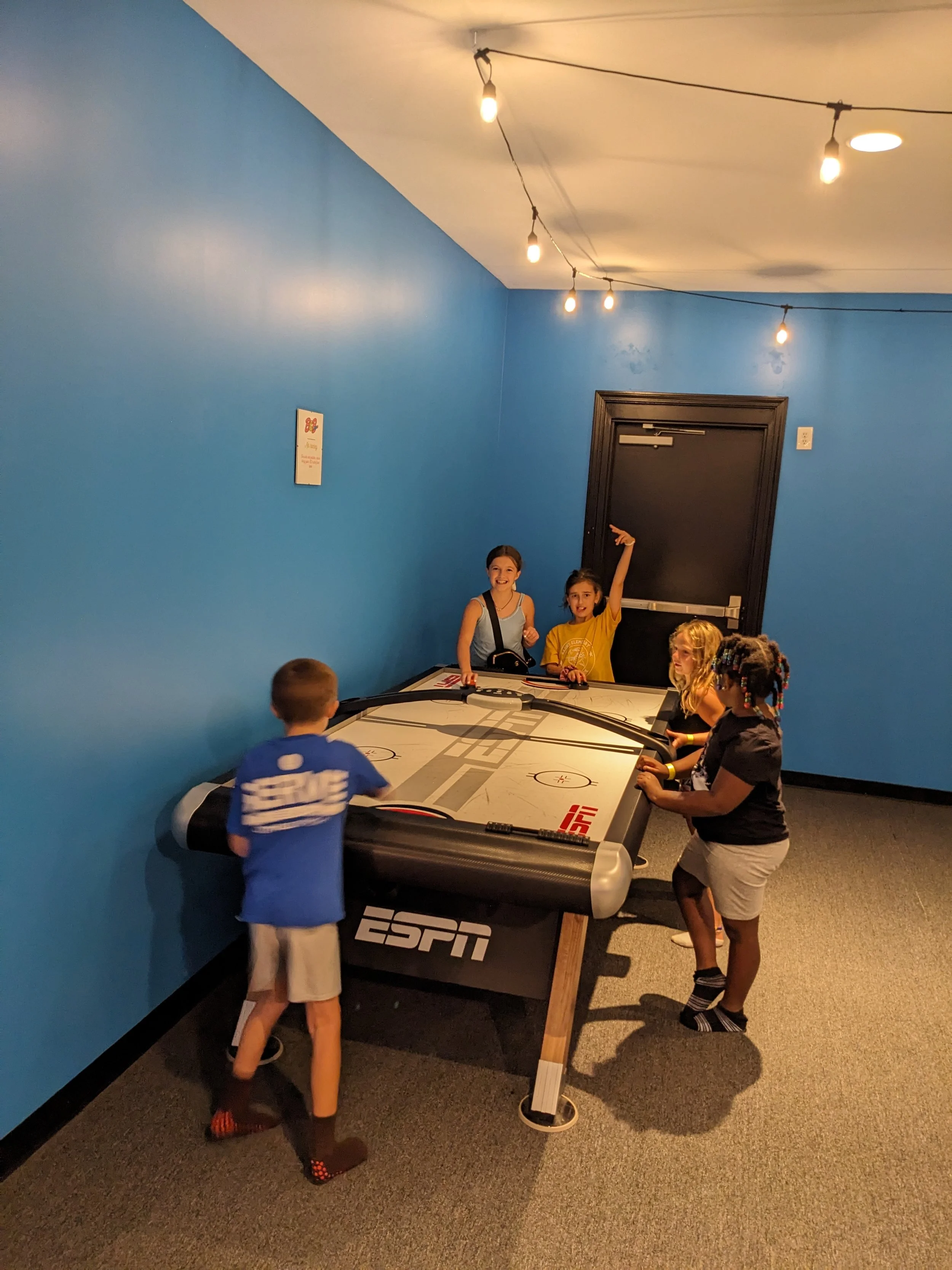 Five children playing air hockey in a room with bright blue walls and string lights hanging from the ceiling.