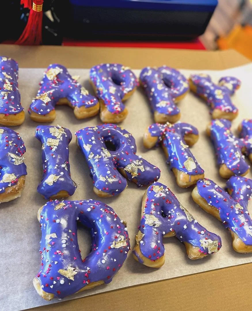 Letter-shaped donuts decorated with purple icing, colorful sprinkles, and gold leaf accents, spelling out "HAPPY DAY" on parchment paper.