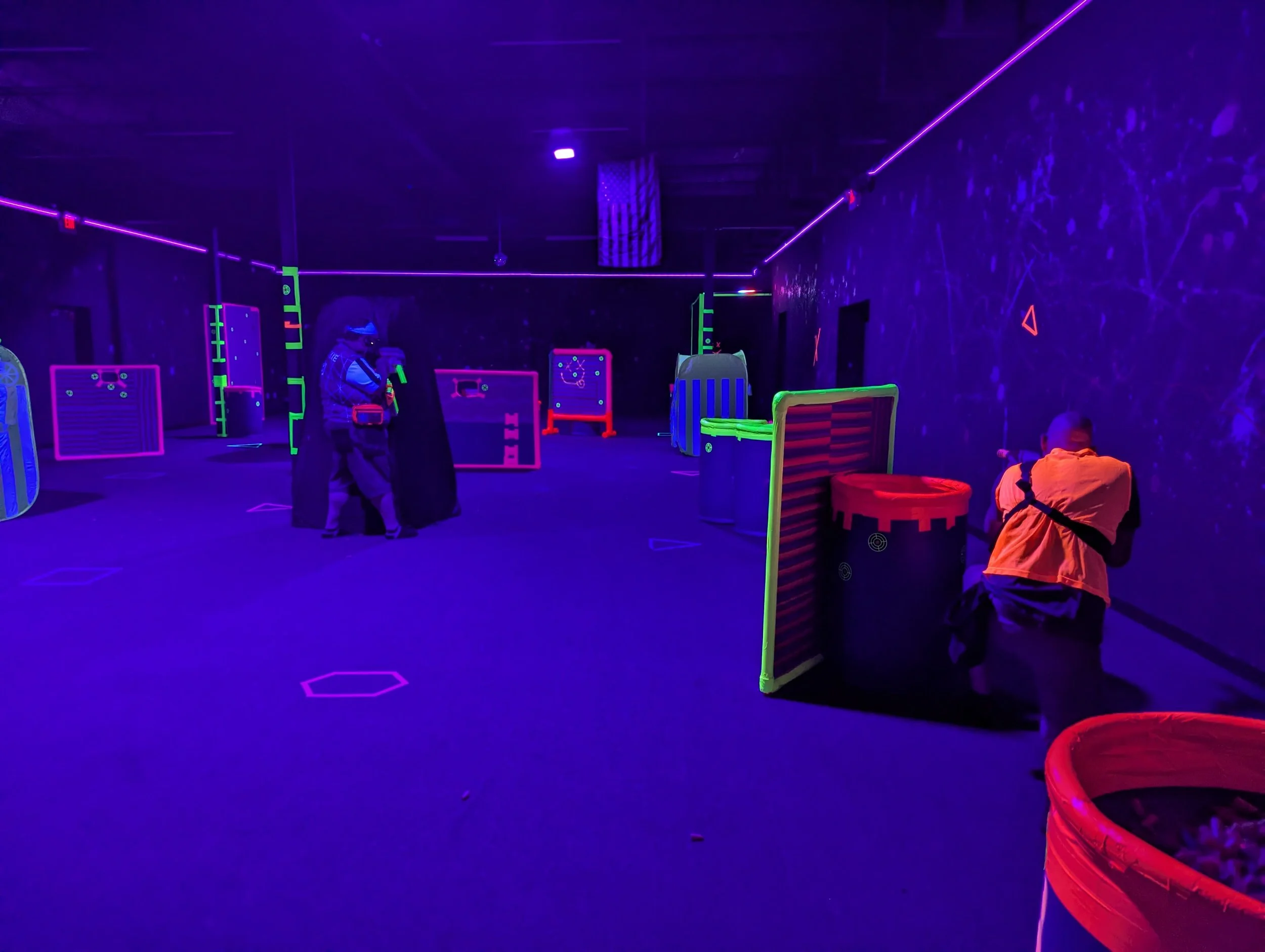 Laser tag arena with neon lights, laser beams, and players hiding behind colorful barriers.