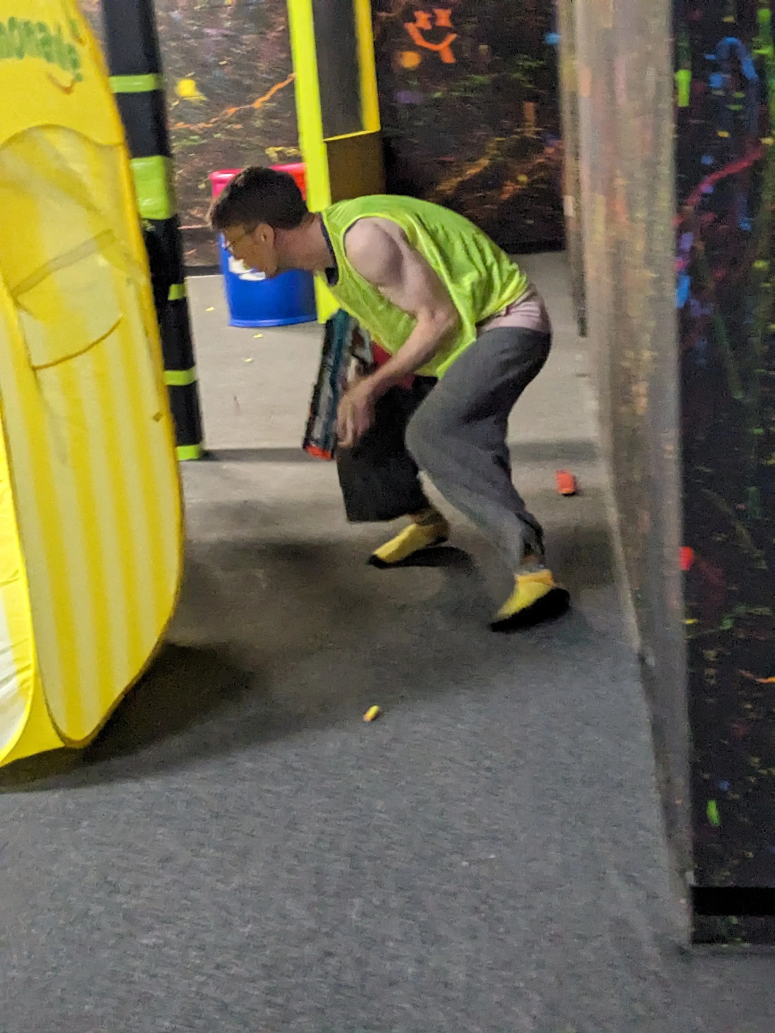 A young man wearing a yellow vest, gray pants, and yellow shoes is crouching or squatting while holding a weapon in an nerf arena.