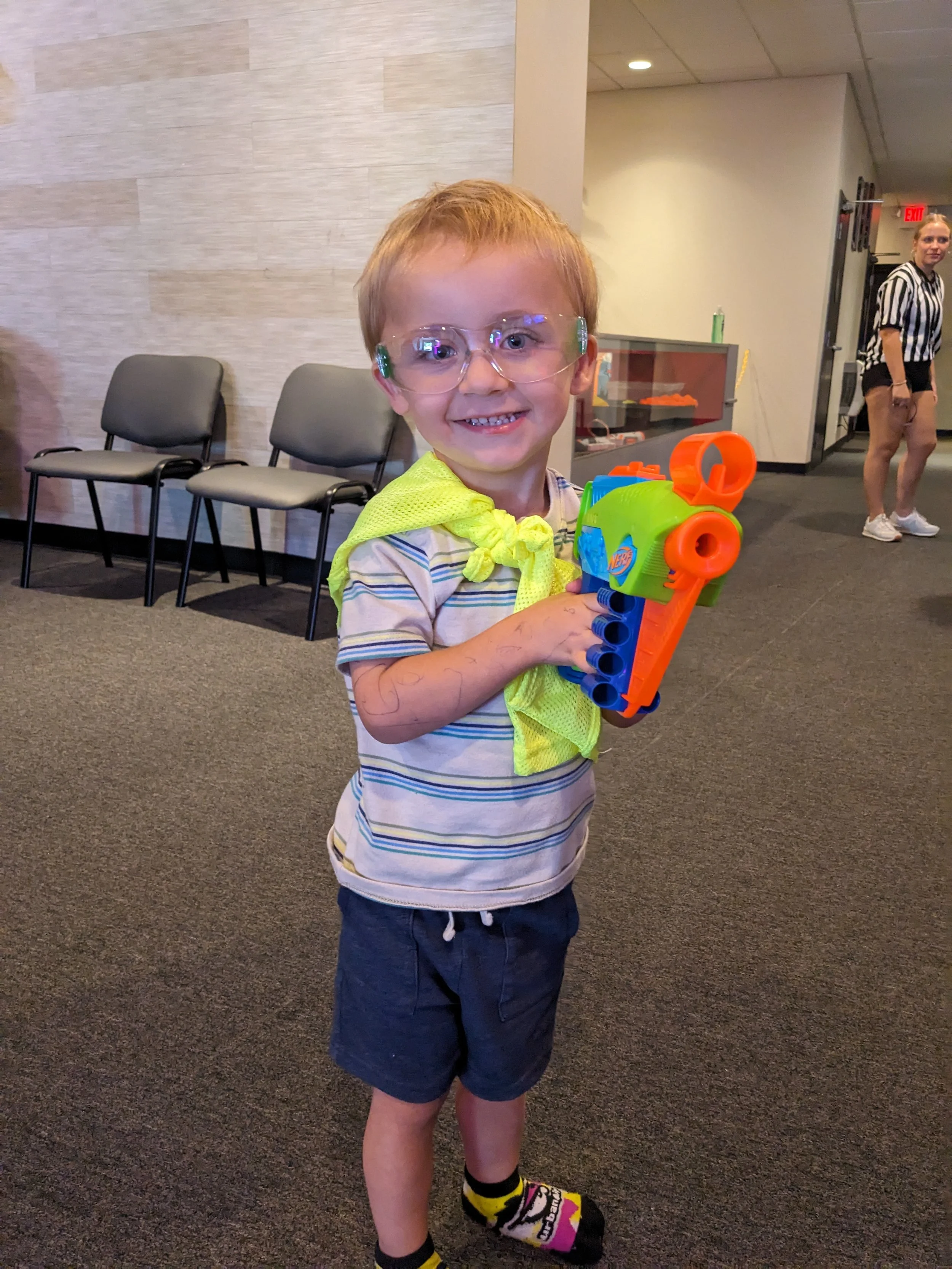 A young boy with glasses and a bright yellow shirt tied around his neck, holding a colorful Nerf gun, smiling at the camera in an indoor setting with chairs and an adult in the background.